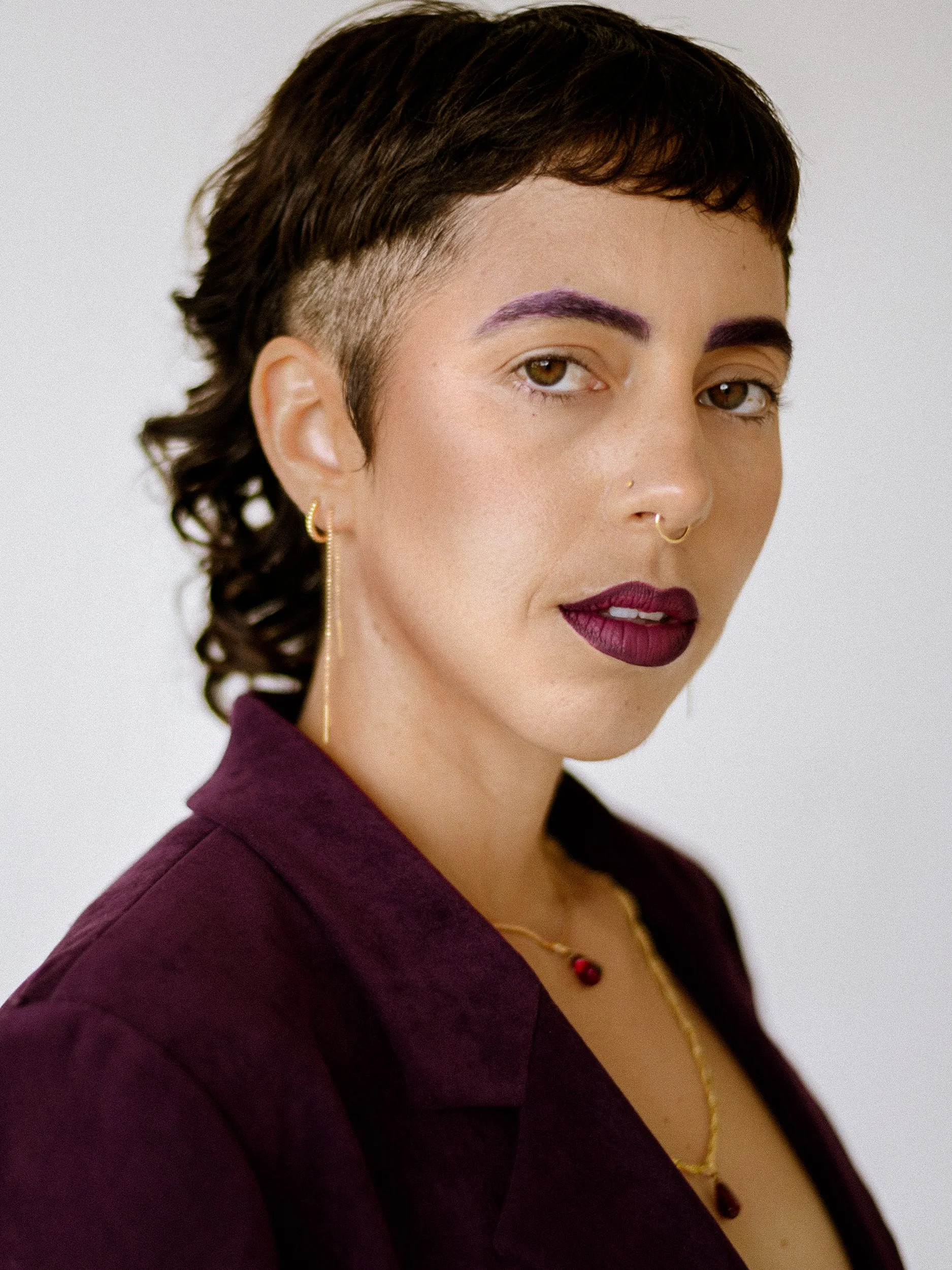 Close-up portrait of a woman with short, dark hair, purple eyebrows, and dark purple lipstick, wearing jewelry including earrings and necklaces, dressed in a dark purple shirt, against a plain white background, photographed during a studio photograph