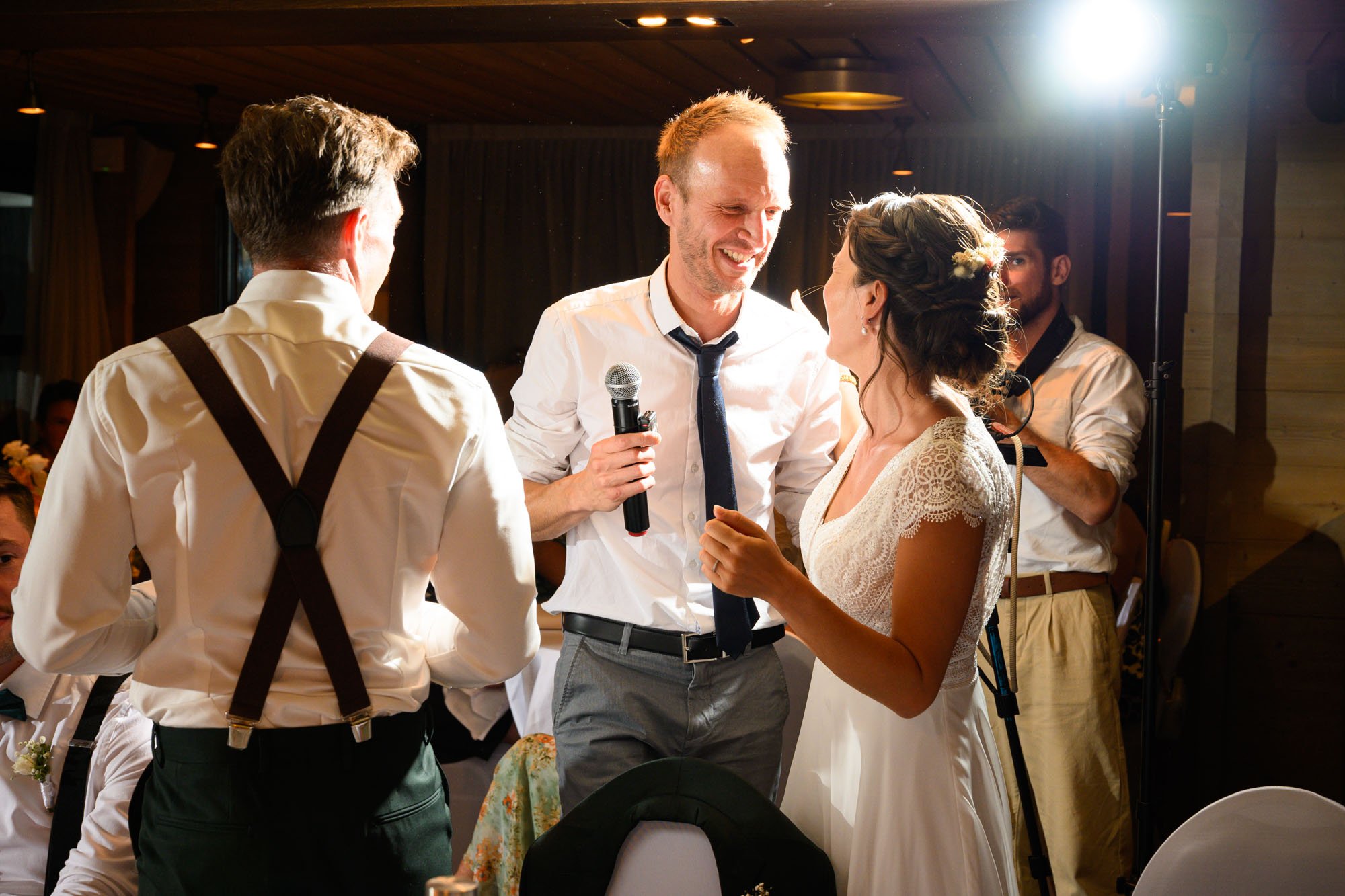 Un couple de mariés échangeant des vœux ou des mots lors de leur mariage, avec un homme tenant un micro, dans une salle de réception chaleureuse.
