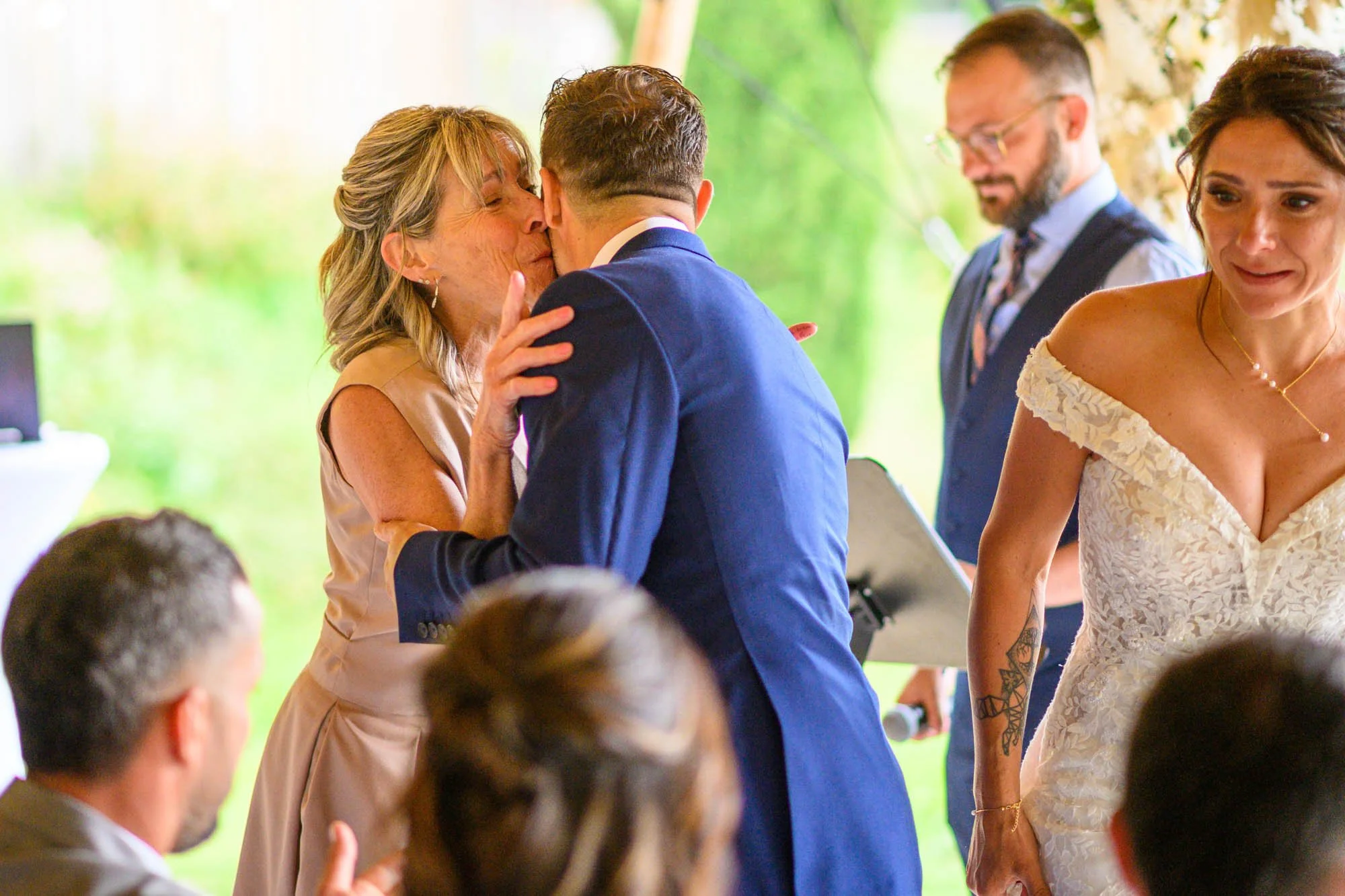Une femme embrasse un homme lors d'une réception de mariage, entourée de plusieurs personnes en tenues élégantes dans un décor vert.