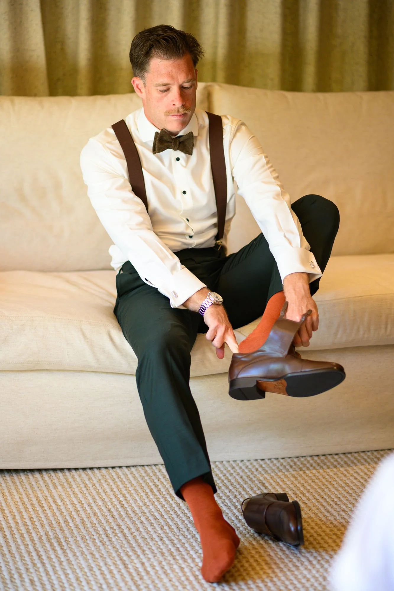 Un homme habillé en smoking, assis sur un canapé, en train de chausser une botte en cuir marron, portant des chaussettes orange, avec une montre à son poignet gauche.