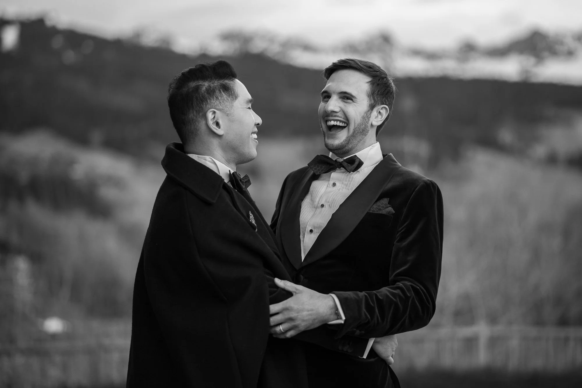 Deux hommes habillés en smoking, souriants et se tenant par la taille, lors d'une cérémonie ou d'un rassemblement en plein air.