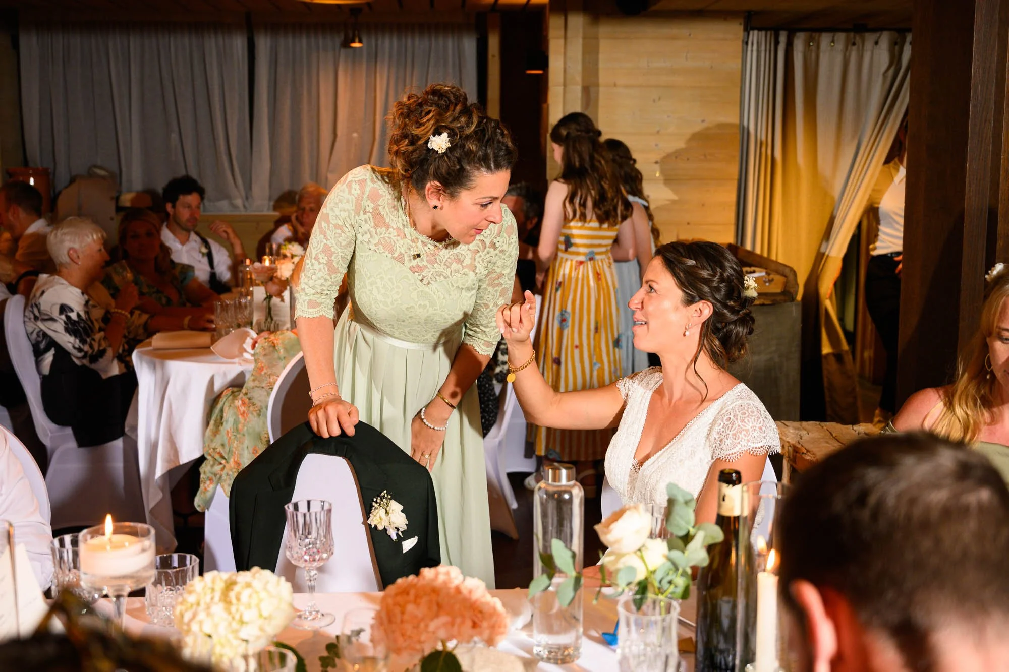 Deux femmes lors d'un mariage, l'une en robe blanche et l'autre en robe vert pâle, partagent un moment de complicité à une table décorée de fleurs et de bougies.