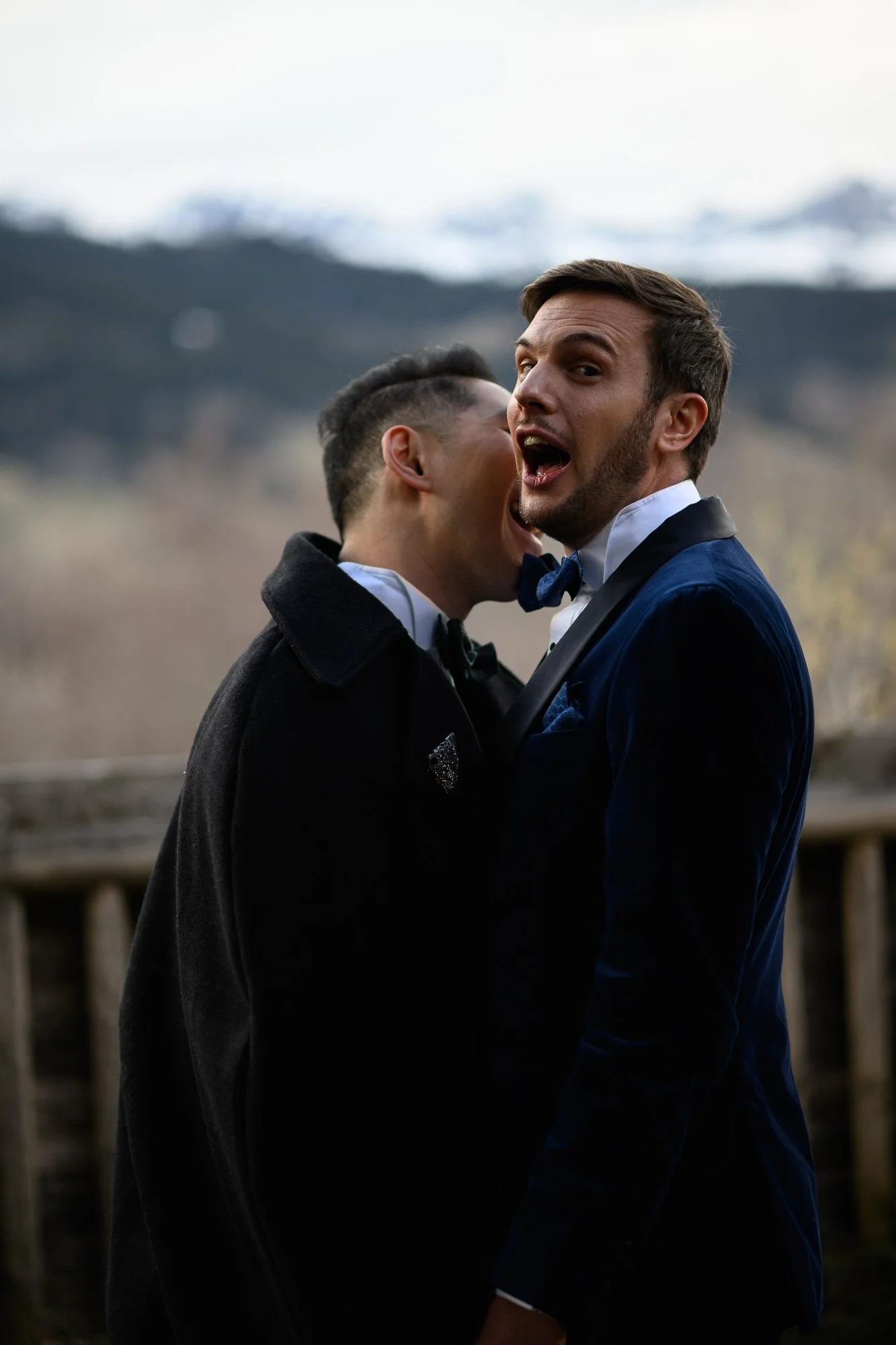 Deux hommes en costumes élégants sont ensemble à l'extérieur avec un arrière-plan de montagnes et de nature.