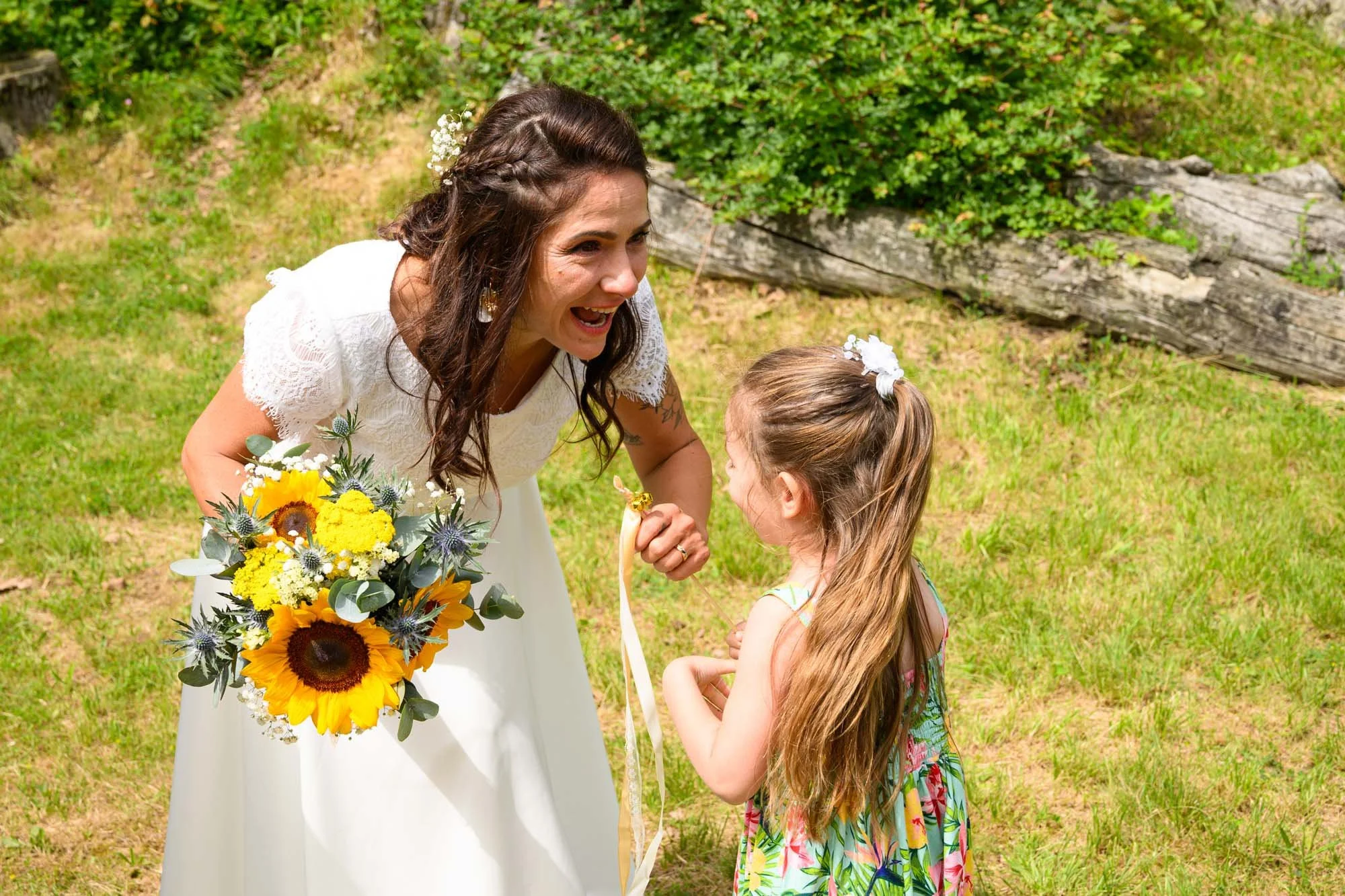Une femme en robe blanche de mariage sourit en regardant une jeune fille lors d'une cérémonie en plein air, avec un bouquet de fleurs en main.