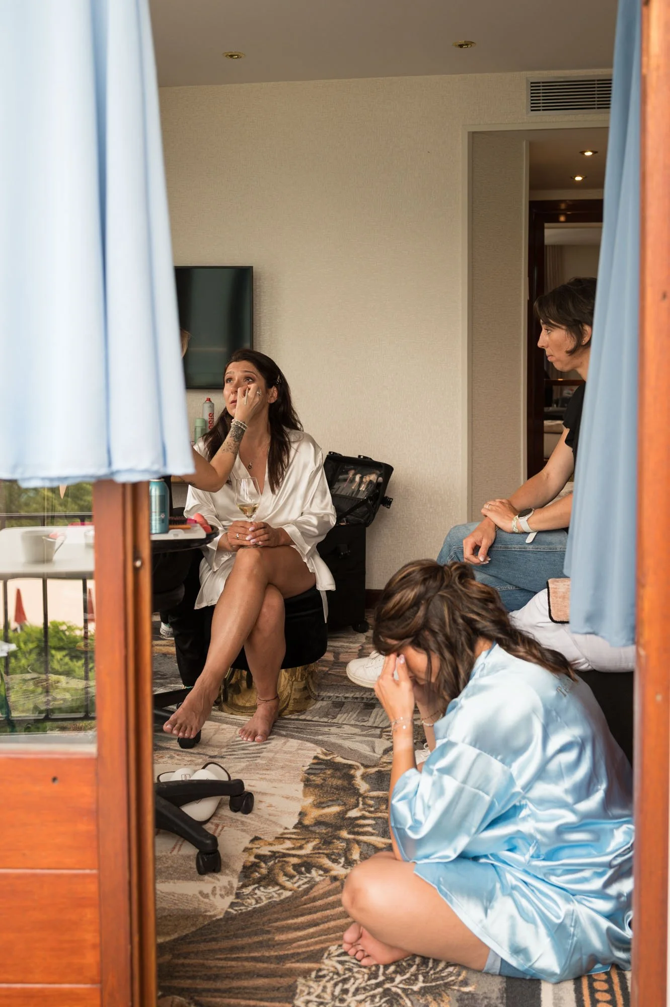 Trois femmes, dont deux en robe de satin, dans une chambre d'hôtel, avec une femme assise sur une chaise tenant un verre de vin, semblant émue ou stressée, et deux autres femmes, l'une assise par terre et l'autre assise sur un lit, semblent en pleurs