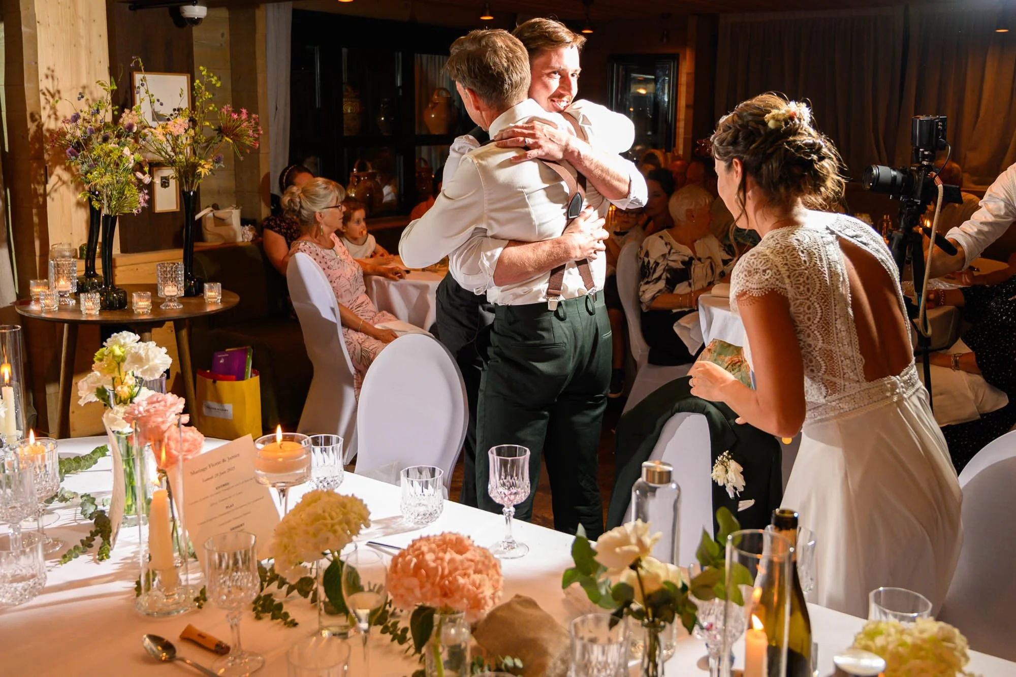 Deux hommes s'embrassent lors d'un mariage, entourés de plusieurs invités assis à des tables décorées, dans un cadre chaleureux avec des fleurs et des bougies.