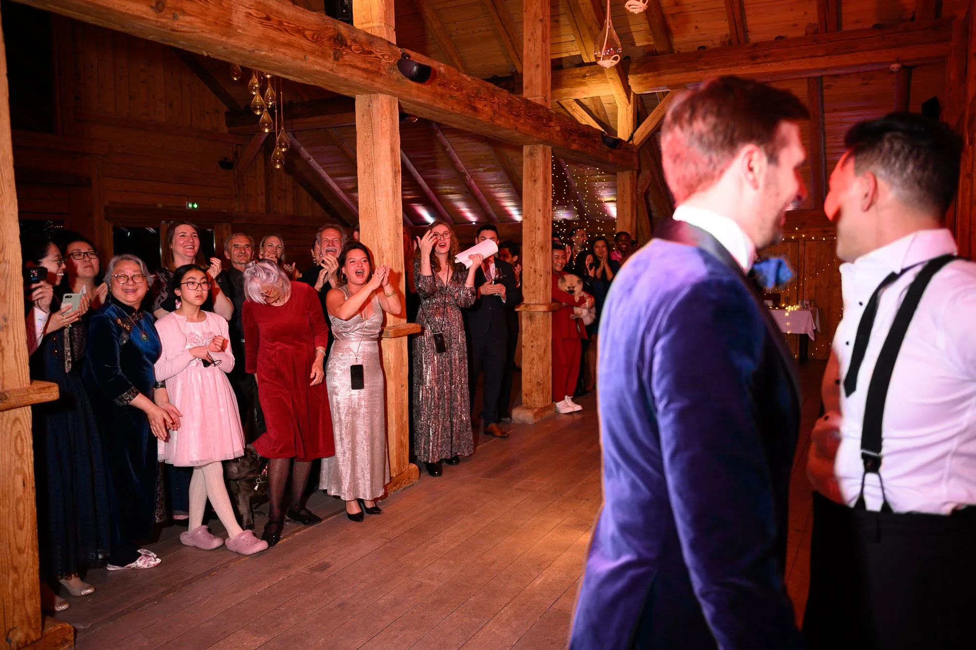 Deux hommes vêtus de smokings entourés de personnes qui sourient et applaudissent lors d'une célébration dans une salle en bois.