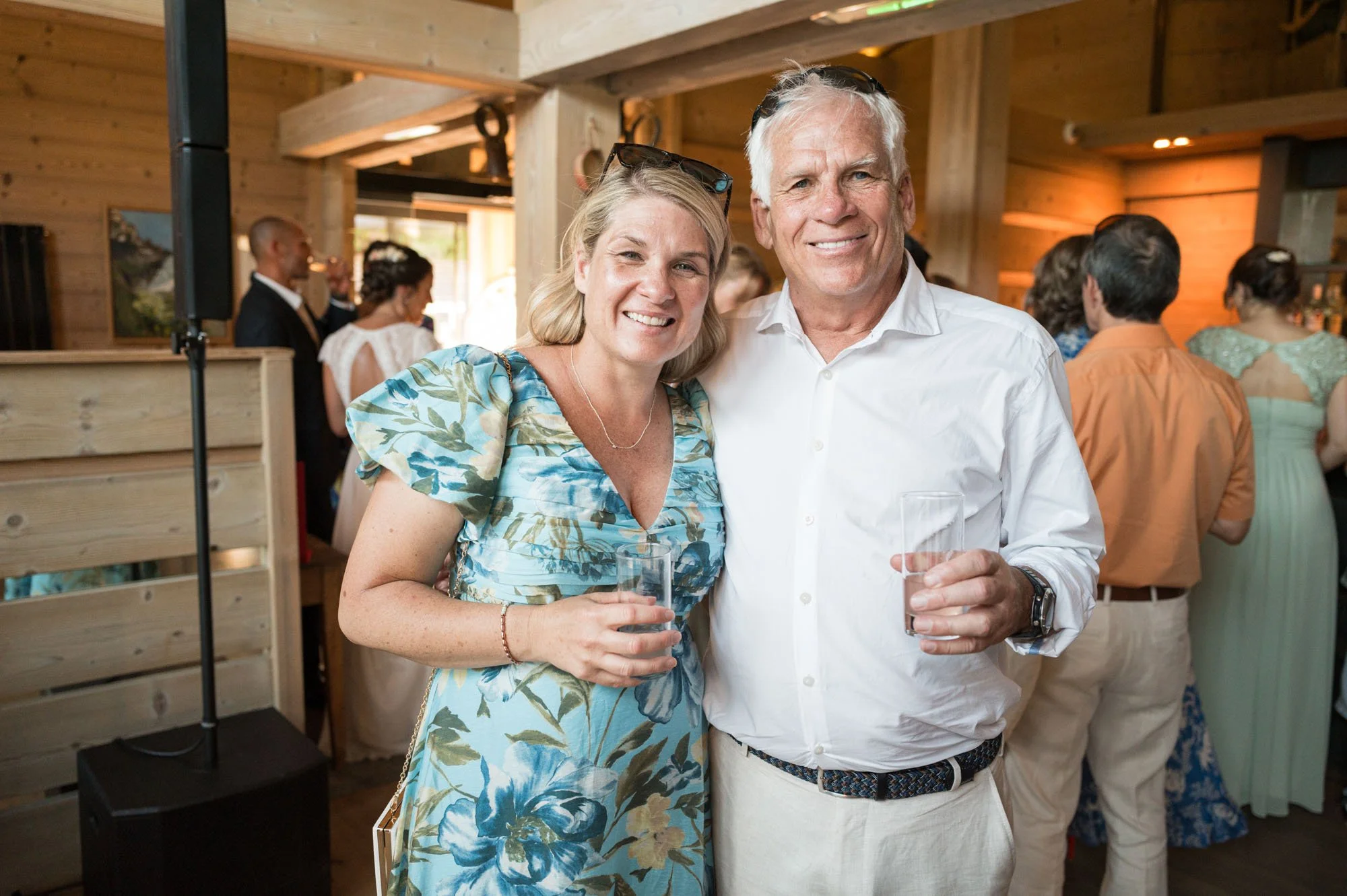 Deux personnes souriantes tenant des verres à cocktail lors d'une fête dans un intérieur en bois avec d'autres invités en arrière-plan.