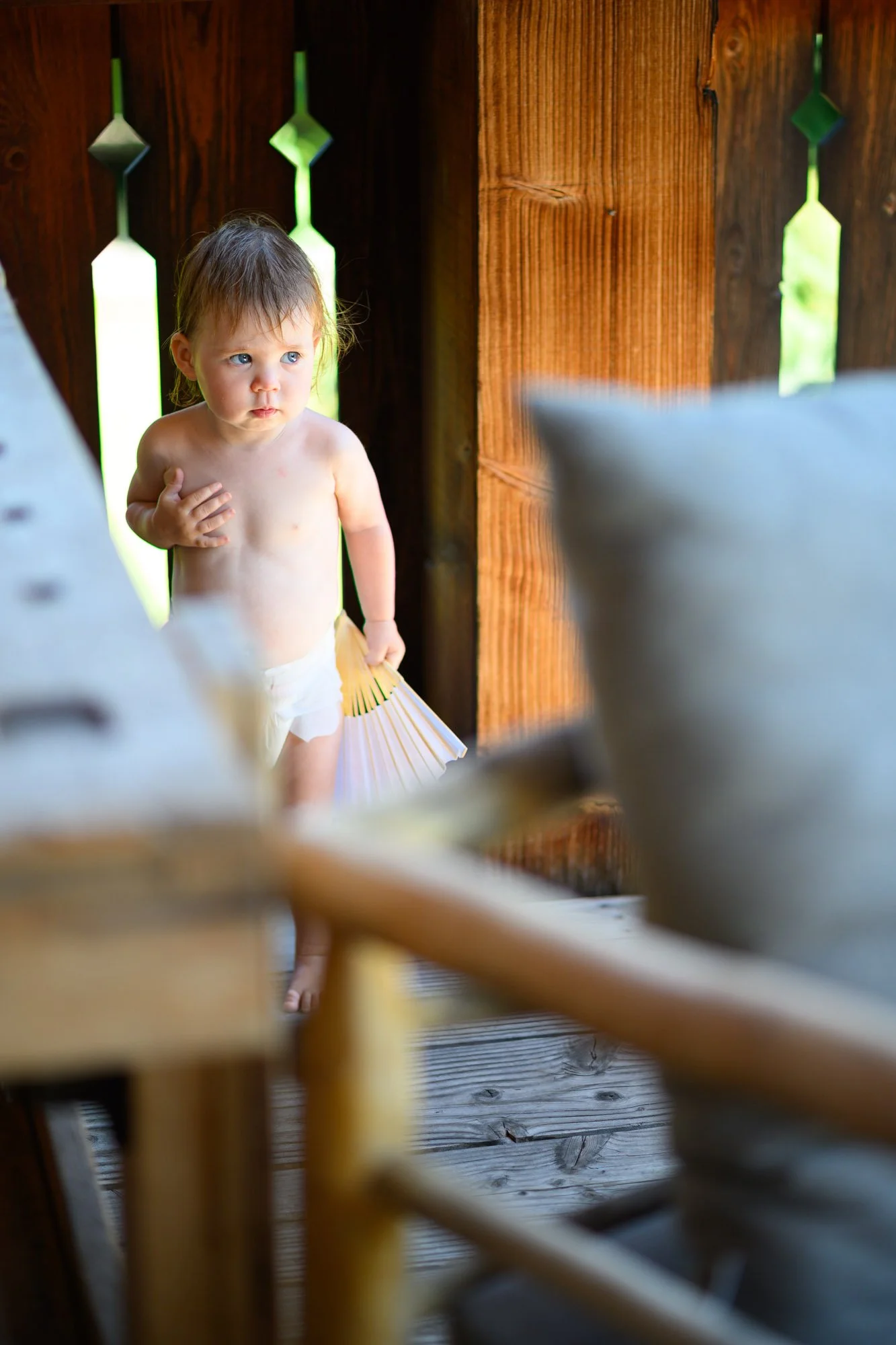 Un jeune enfant debout dans une cabane en bois, partiellement vêtu, tenant un éventail en papier, regardant avec curiosité ou surprise.