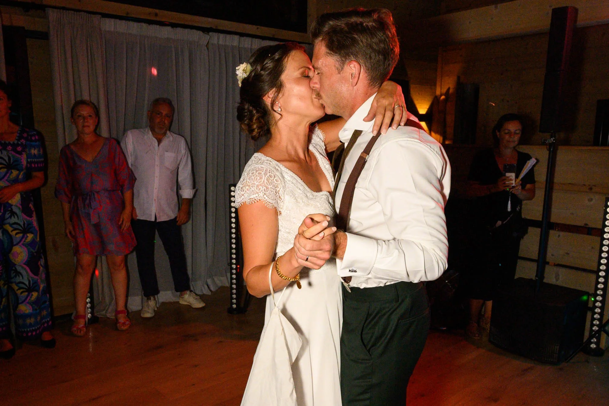 Un couple danse lors d'un mariage, eux, portant une robe blanche et un costume blanc avec une cravate noire, s'embrassent alors qu'ils dansent, entourés d'invités.