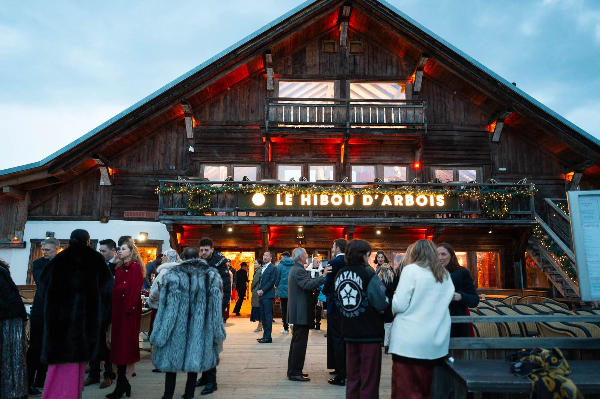 Une foule de personnes devant un bâtiment en bois avec un panneau lumineux indiquant "Le Hibou d'Arbois", décoré de guirlandes lumineuses, en soirée.