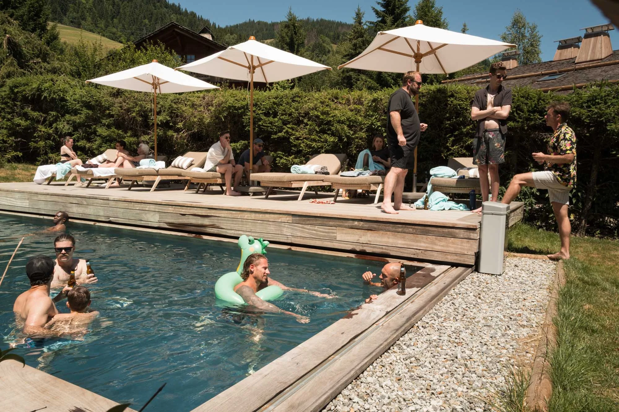 Groupe de personnes profitant d'une journée ensoleillée au bord d'une piscine en pleine nature, avec des chaises longues, parasols, et un environnement verdoyant.