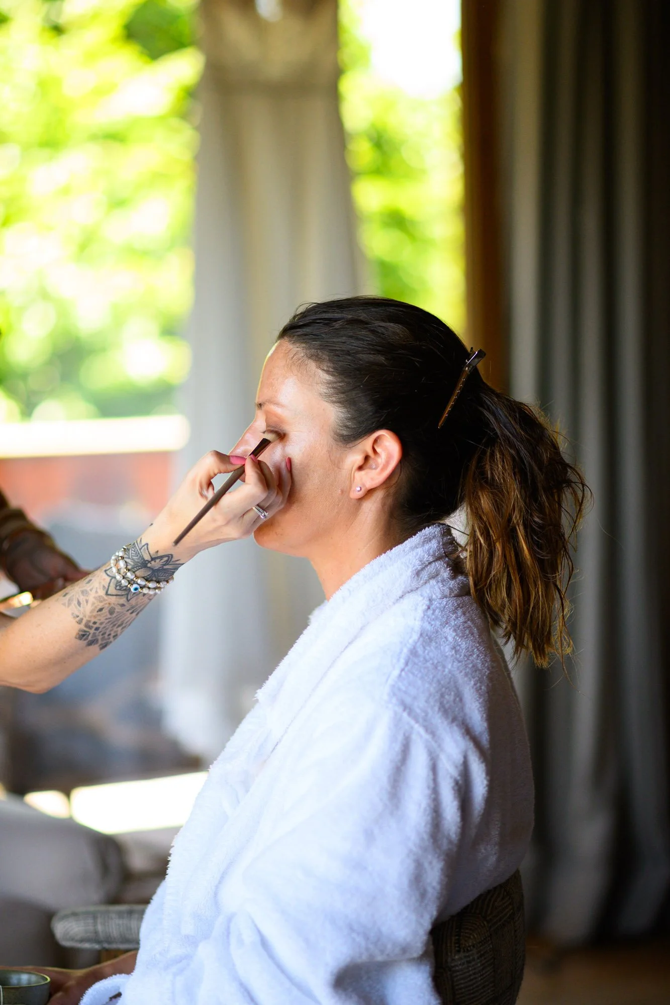Une femme reçoit un maquillage lors d'une séance de maquillage, elle porte une robe blanche et est assise devant une fenêtre avec une lumière naturelle.
