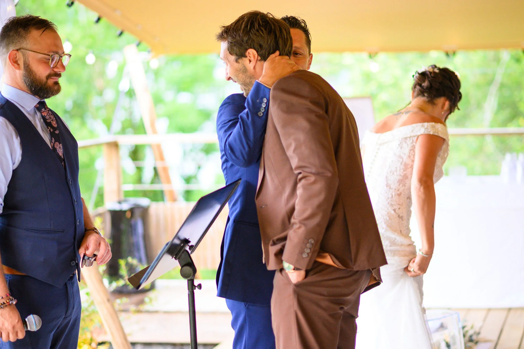 Homme en costume bleu embrassant un autre homme en costume marron lors d'une cérémonie de mariage. Une femme en robe blanche se tient à l'arrière, légèrement inclinée. Un homme avec une veste bleue et une cravate à motifs se tient à gauche, avec un m