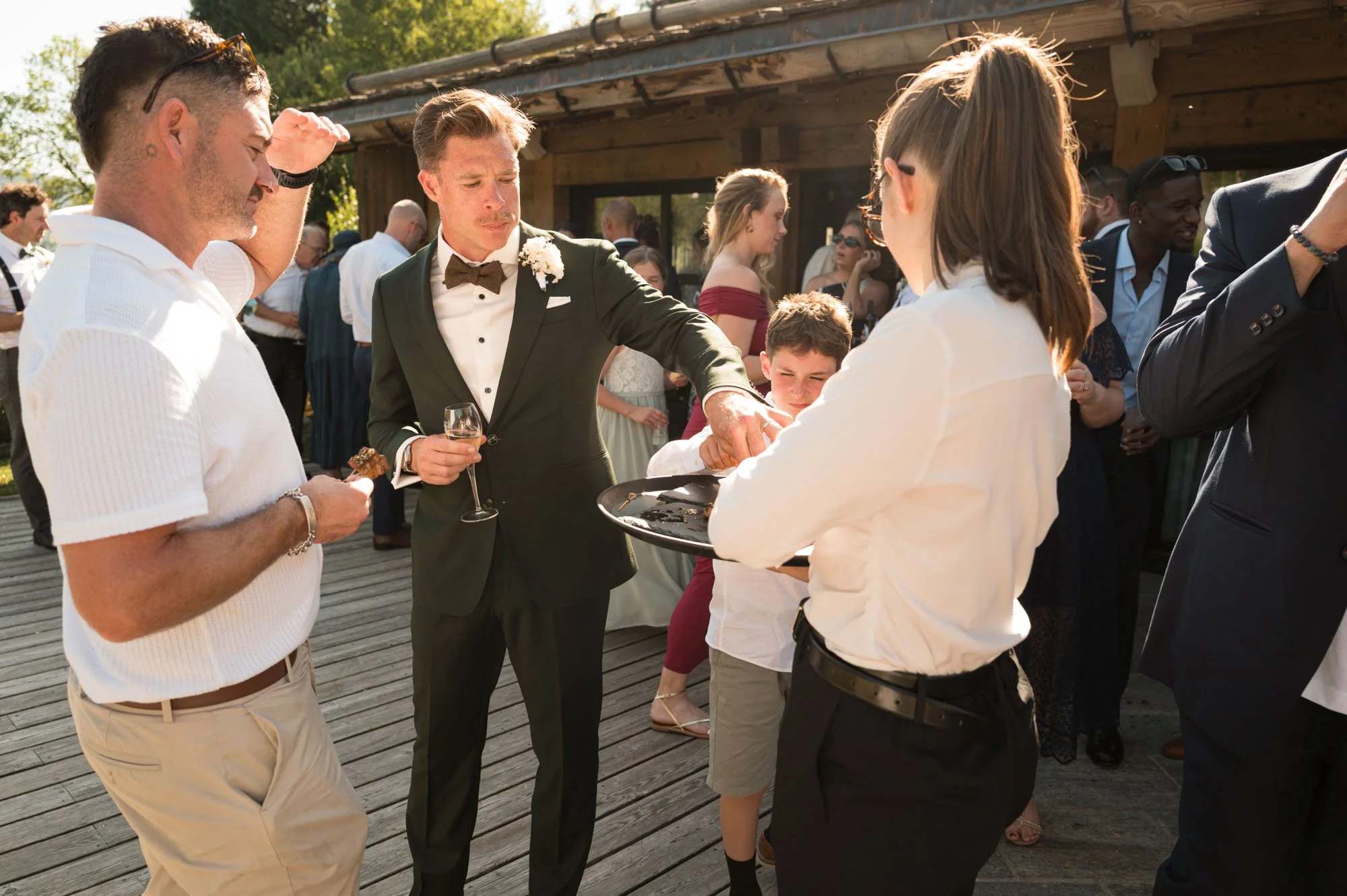 Groupe de personnes lors d'une fête ou d'un mariage en extérieur, avec un homme en costume noir avec un nœud papillon, une femme avec un plateau de service, et d'autres personnes qui socialisent sous un ciel ensoleillé.