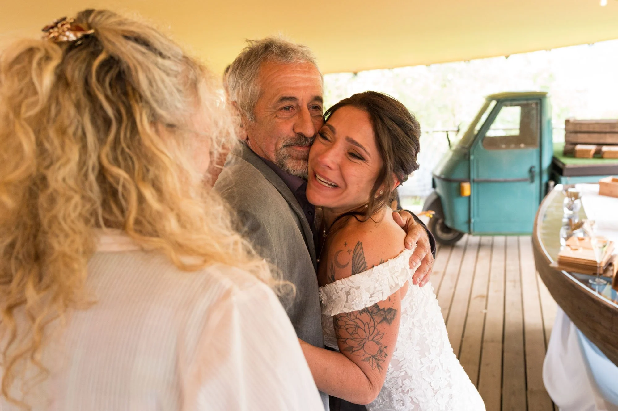 Une mariée en robe de mariage embrasse un homme lors d'une cérémonie de mariage, une femme aux cheveux blonds bouclés assiste. En arrière-plan, un véhicule bleu et une table avec des objets.