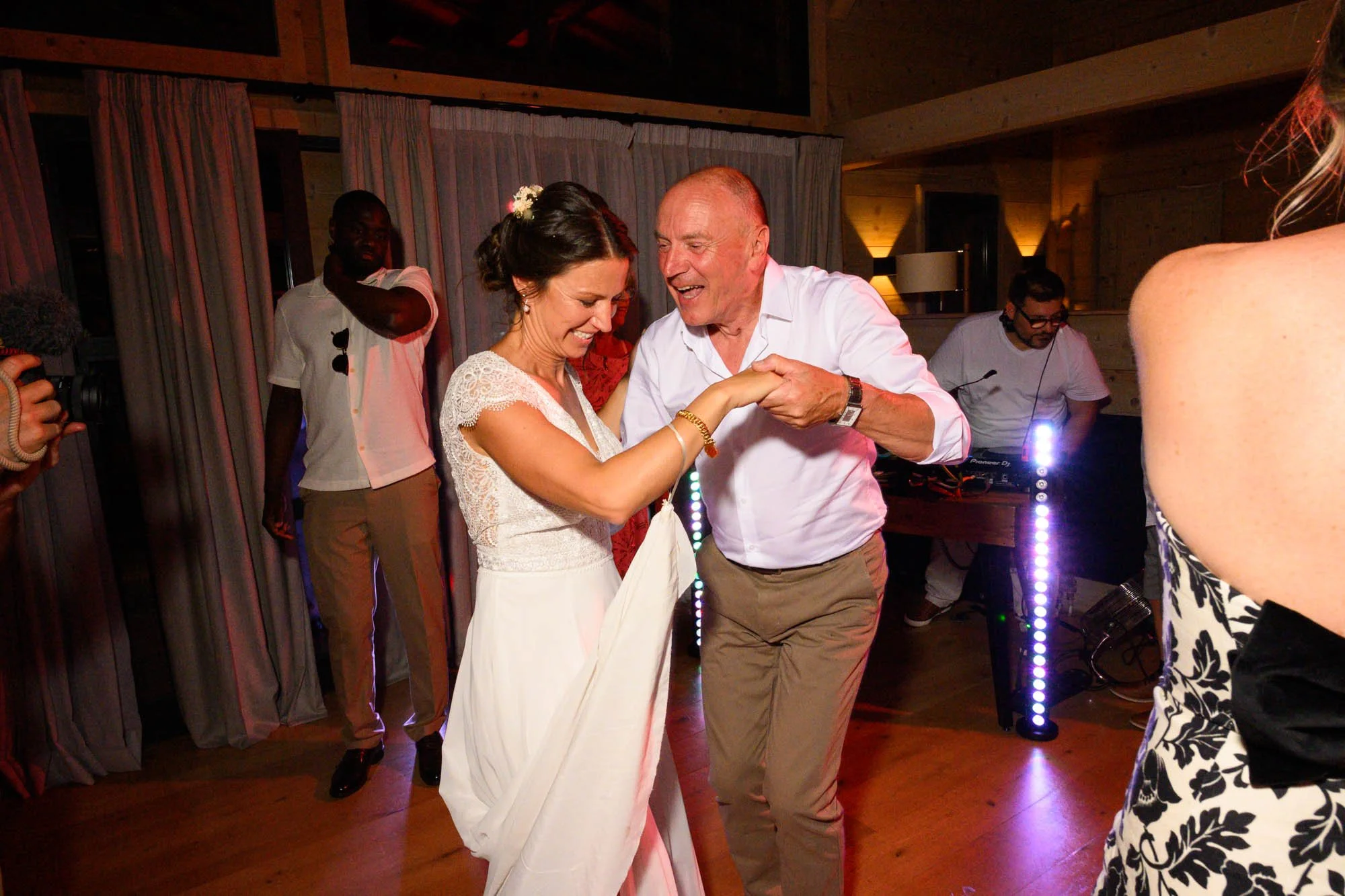 Deux personnes dansent lors d'un mariage, une femme en robe blanche et un homme en chemise blanche, avec un DJ qui joue de la musique en arrière-plan.