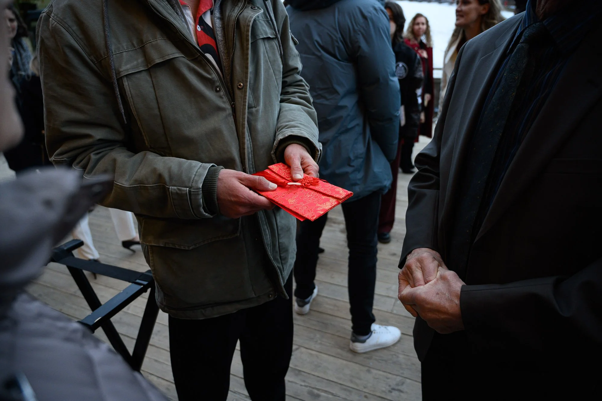 Une personne remet un paquet cadeau rouge à une autre personne lors d'une rencontre en plein air, entourée d'autres personnes.