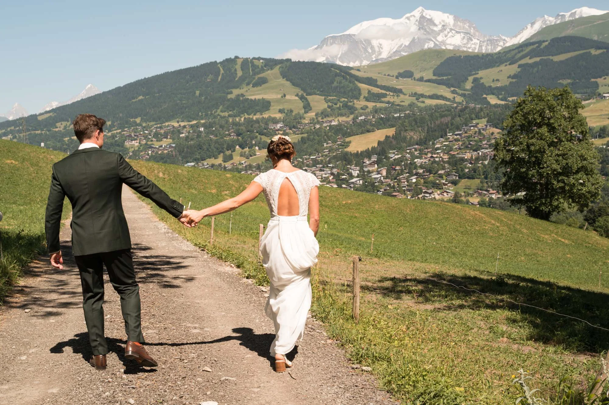 Un couple habillé en mariage se tenant la main en marchant sur un chemin rural avec un paysage de montagnes et de collines verdoyantes en arrière-plan.