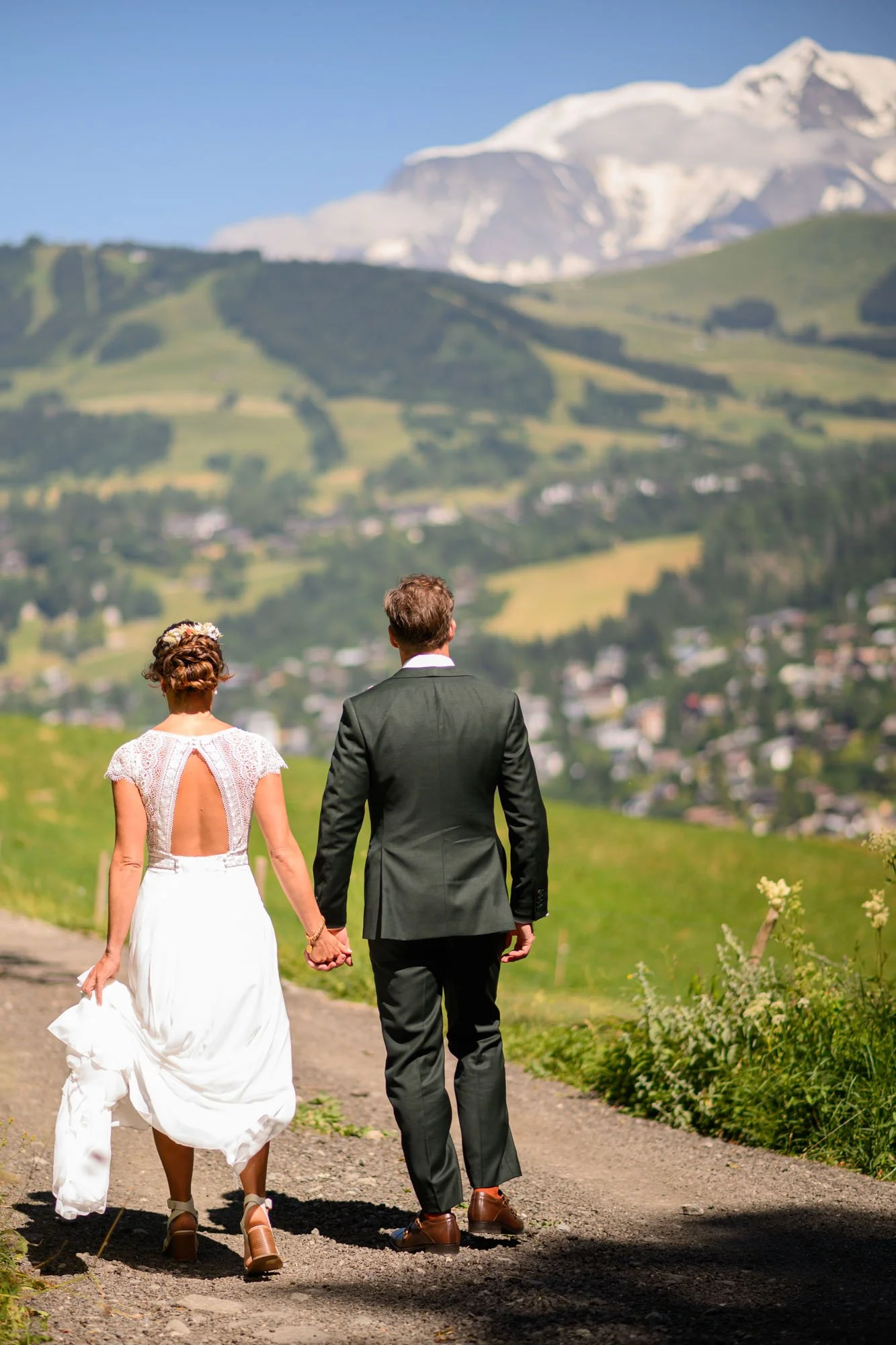 Un couple de mariés marche main dans la main sur un chemin de campagne, vue sur des montagnes enneigées.