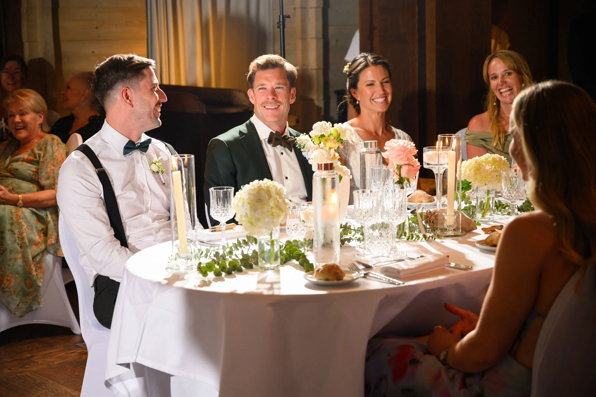 Groupe de personnes souriantes lors d'un repas de mariage, assises autour d'une table décorée de fleurs et de chandelles.
