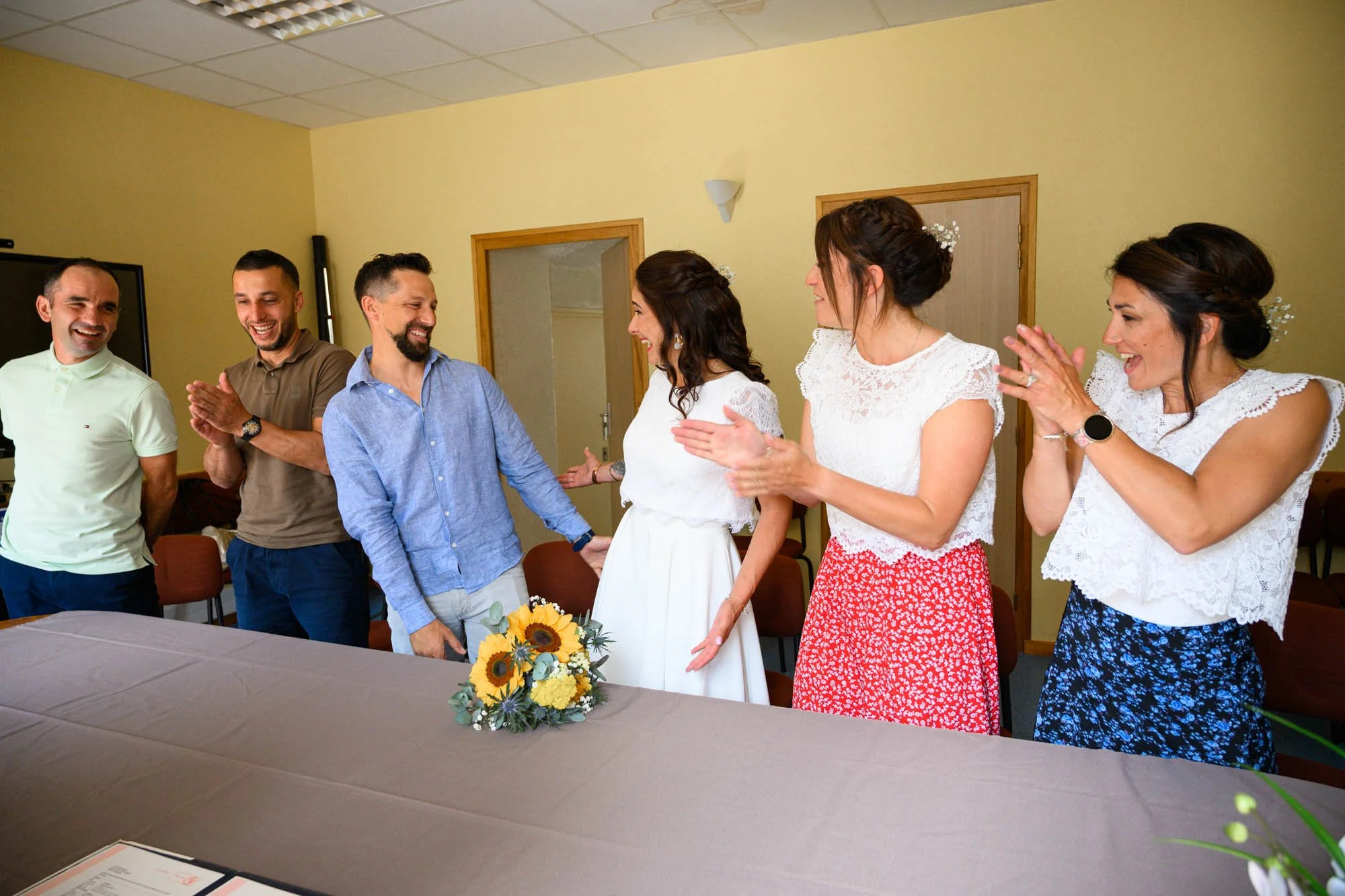 Un groupe de six personnes, dont deux femmes en robe blanche, en train de féliciter et d'applaudir une femme en robe blanche, dans une salle avec une table couverte d'une nappe beige, un bouquet de tournesols, et des chaises.