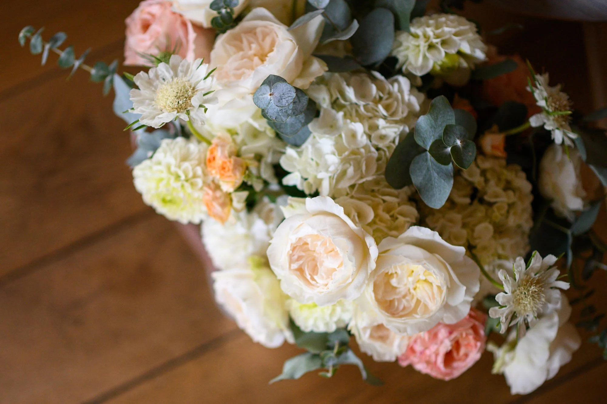 Un bouquet de fleurs composé de roses, hortensias, eucalyptus et autres fleurs blanches et rose pâle sur un fond de sol en bois.