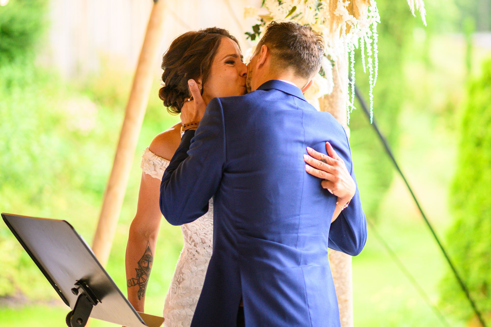 Un couple se embrasse lors de leur mariage en plein air, sous une arche décorée de fleurs, avec un fond de nature verdoyante.