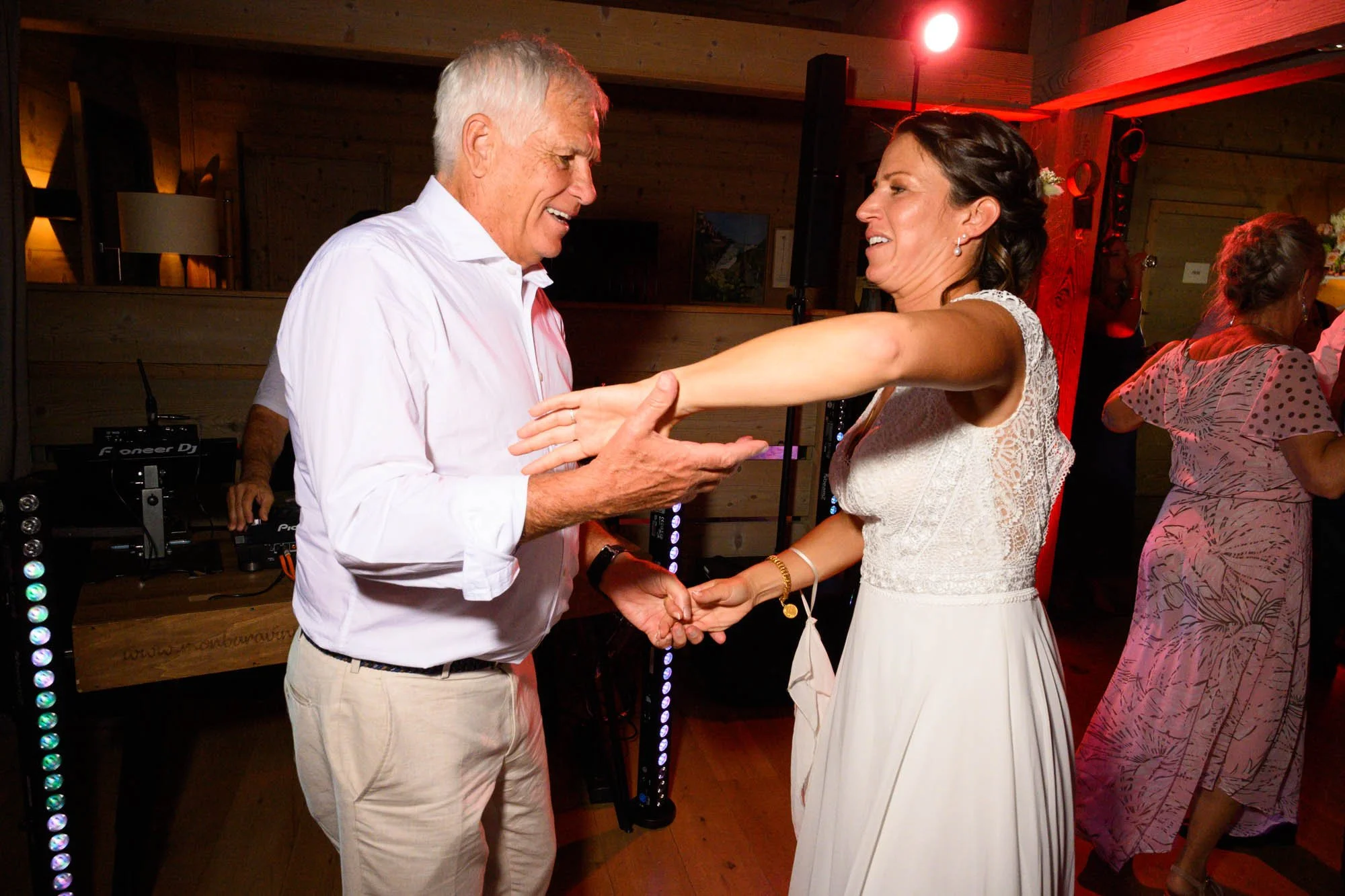 Un couple danse lors d'une célébration, la femme porte une robe blanche en dentelle et l'homme porte une chemise blanche et un pantalon beige, dans une ambiance chaleureuse avec d'autres personnes en arrière-plan.