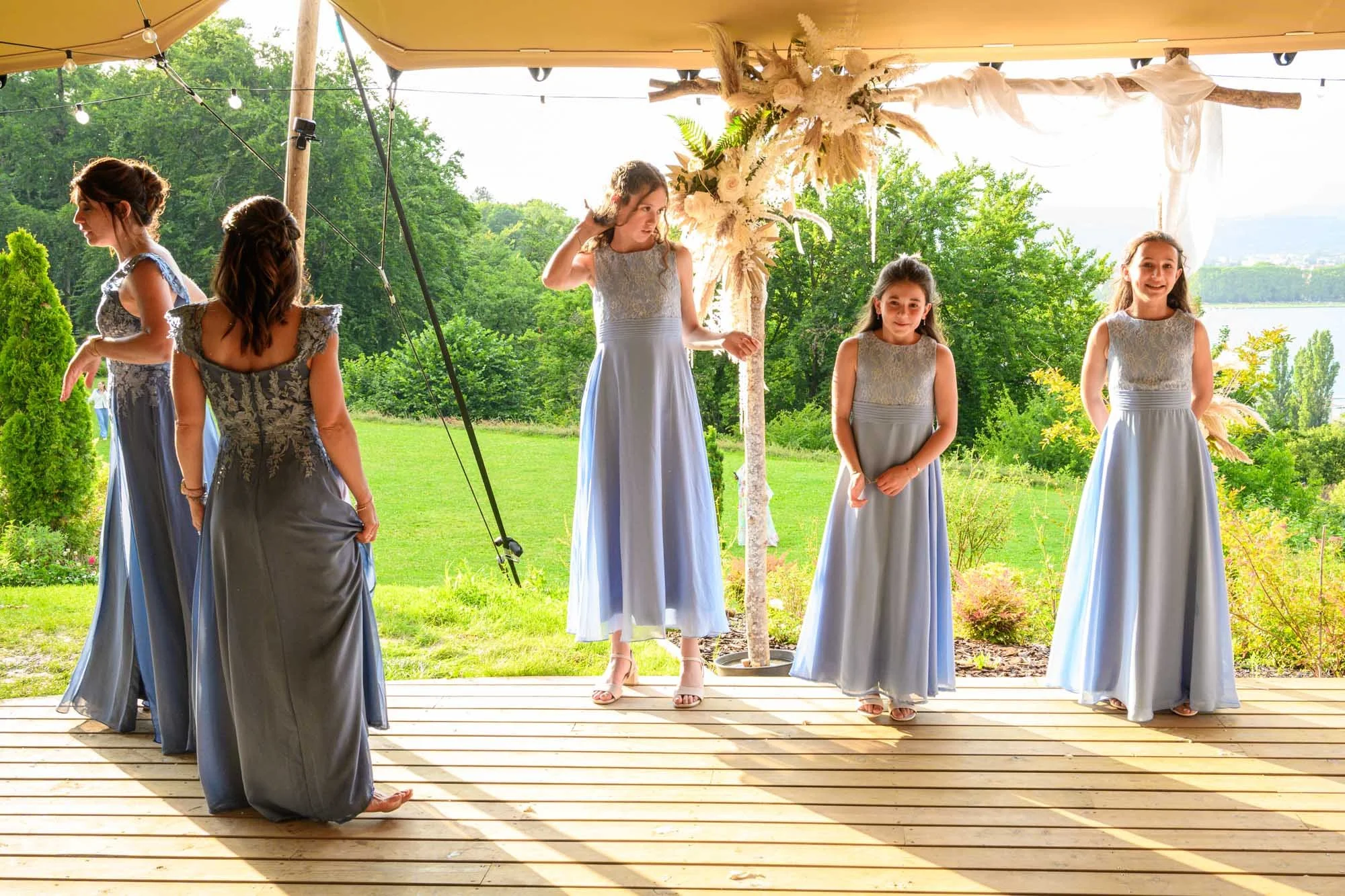 Groupe de jeunes filles en robes bleues longue turquoise, posant lors d'un événement en plein air sous un chapiteau décoré avec des fleurs, avec un paysage verdoyant en arrière-plan.