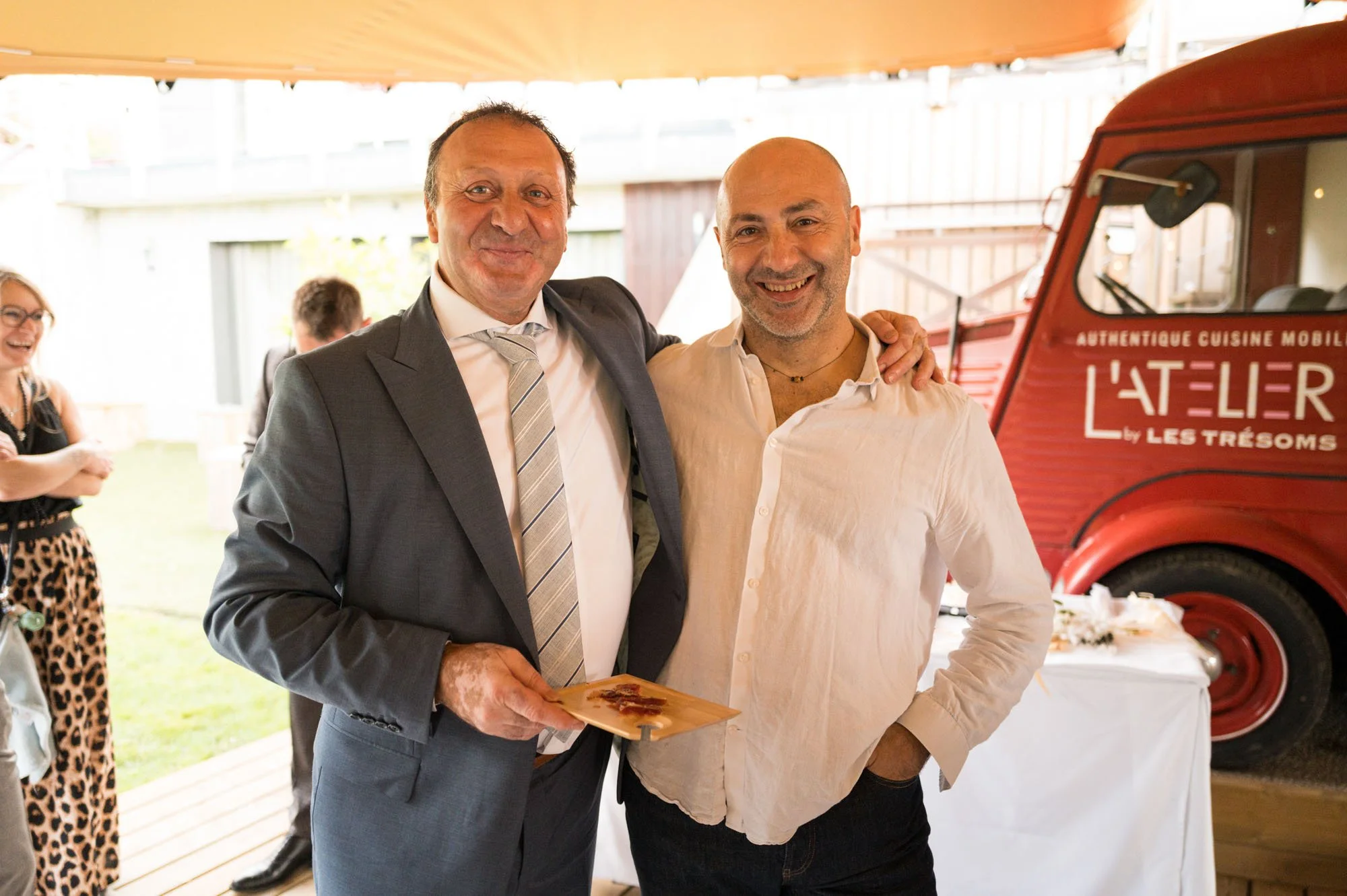 Deux hommes souriants posent ensemble lors d'un événement, avec un camion rouge en arrière-plan portant l'inscription "L'Atelier par Les Trésoms".