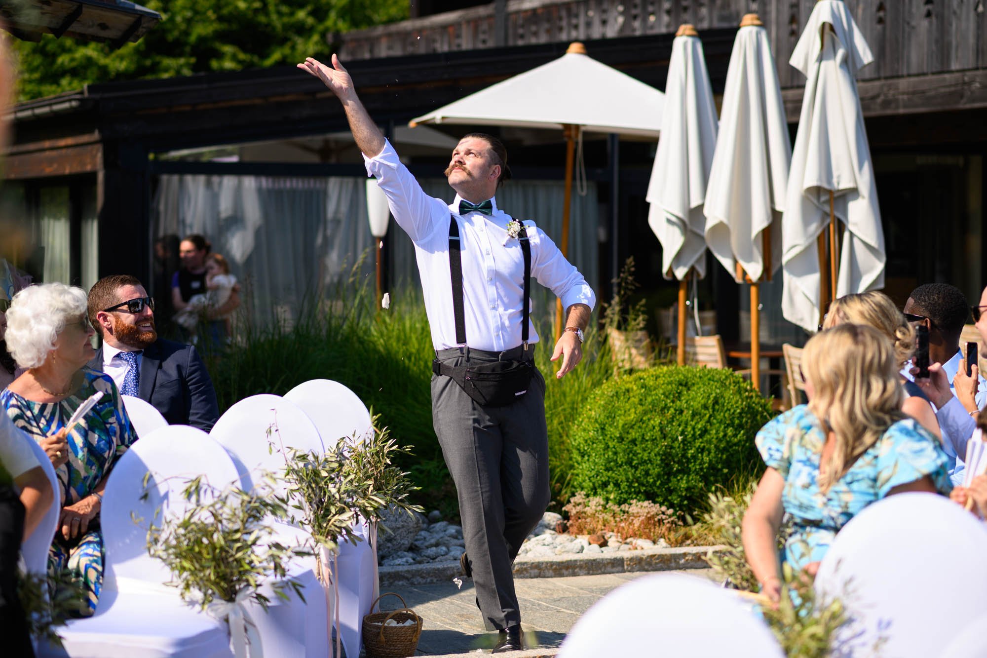 Un homme en costume blanc avec une cravate papillon, faisant une prestation lors d'un mariage en extérieur, entouré d'invités assis.