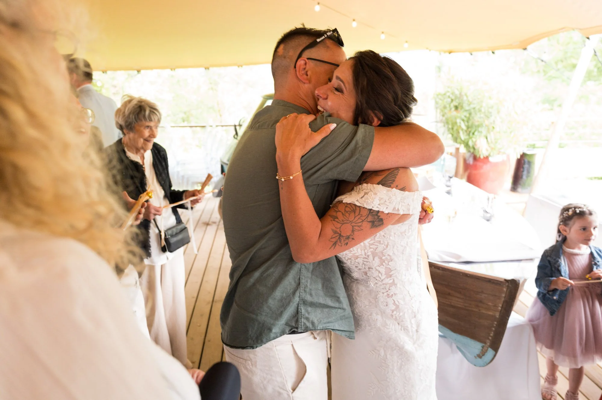 Deux personnes s'embrassent tendrement lors d'une cérémonie, entourées de convives, dans un lieu lumineux avec une décoration naturelle.