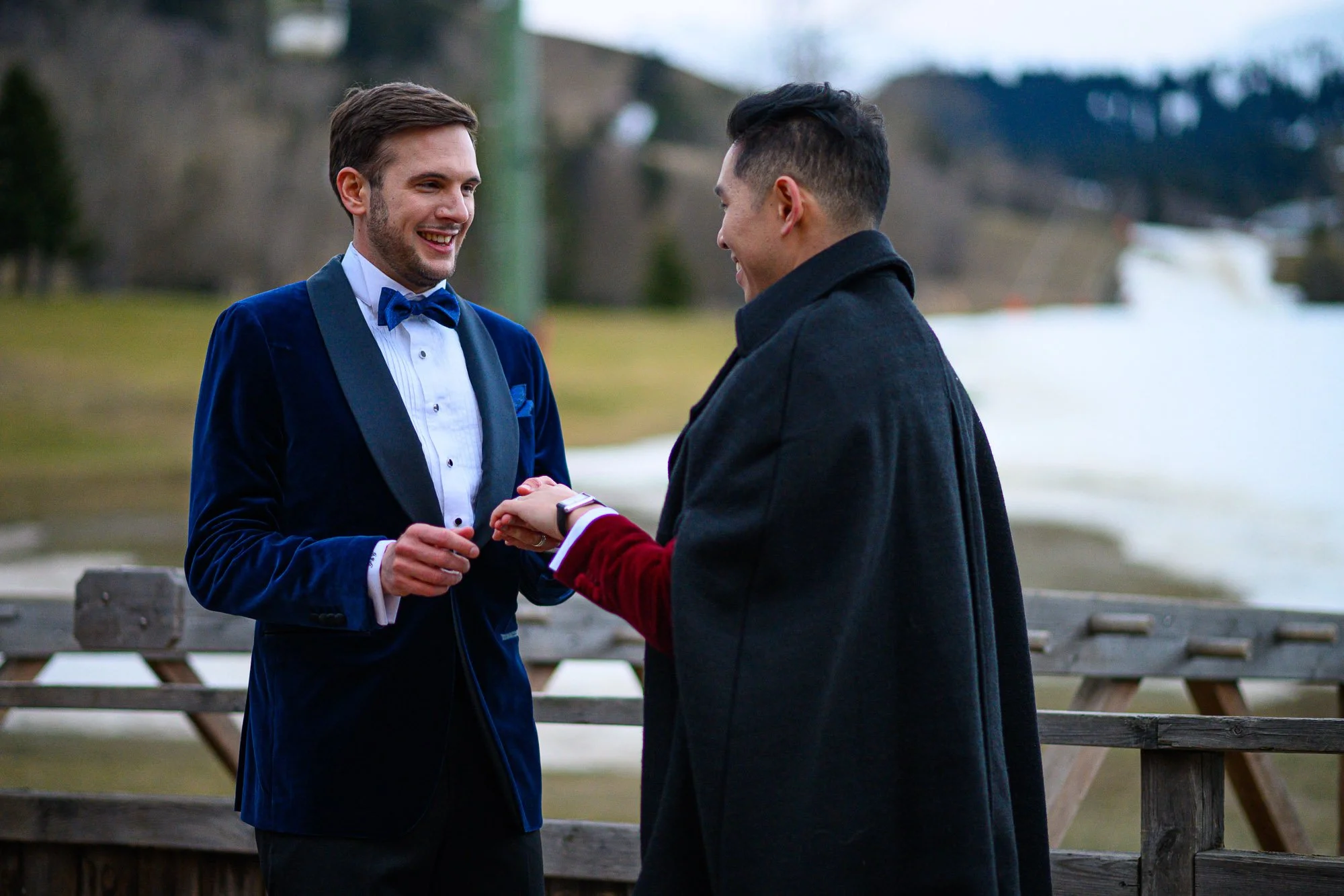 Deux hommes habillés formellement échangent une promesse de mariage en pleine nature près d'un lac, avec un pont en bois en arrière-plan.