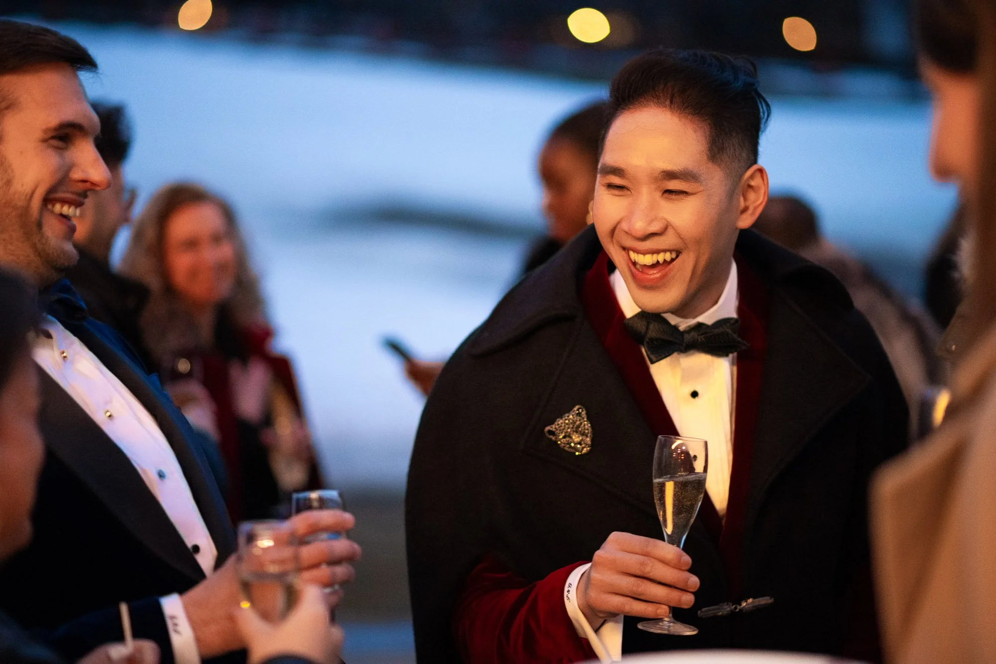 Groupe de personnes habillées élégamment en train de discuter et de boire lors d'une soirée en extérieur près d'une rivière, avec un jeune homme souriant tenant une flûte à champagne.