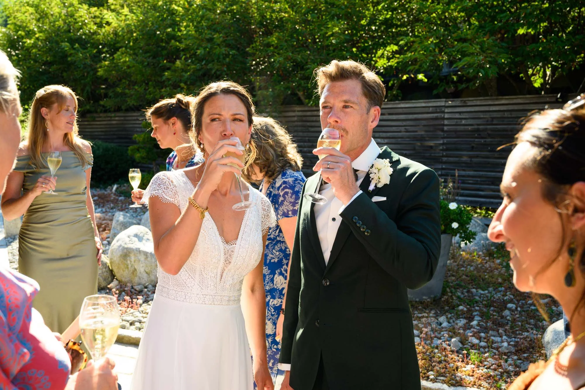 Un groupe de personnes portant des tenues formelles en train de boire du champagne lors d'une fête ou d'un mariage en plein air, avec un fond de végétation verte et un mur en bois.