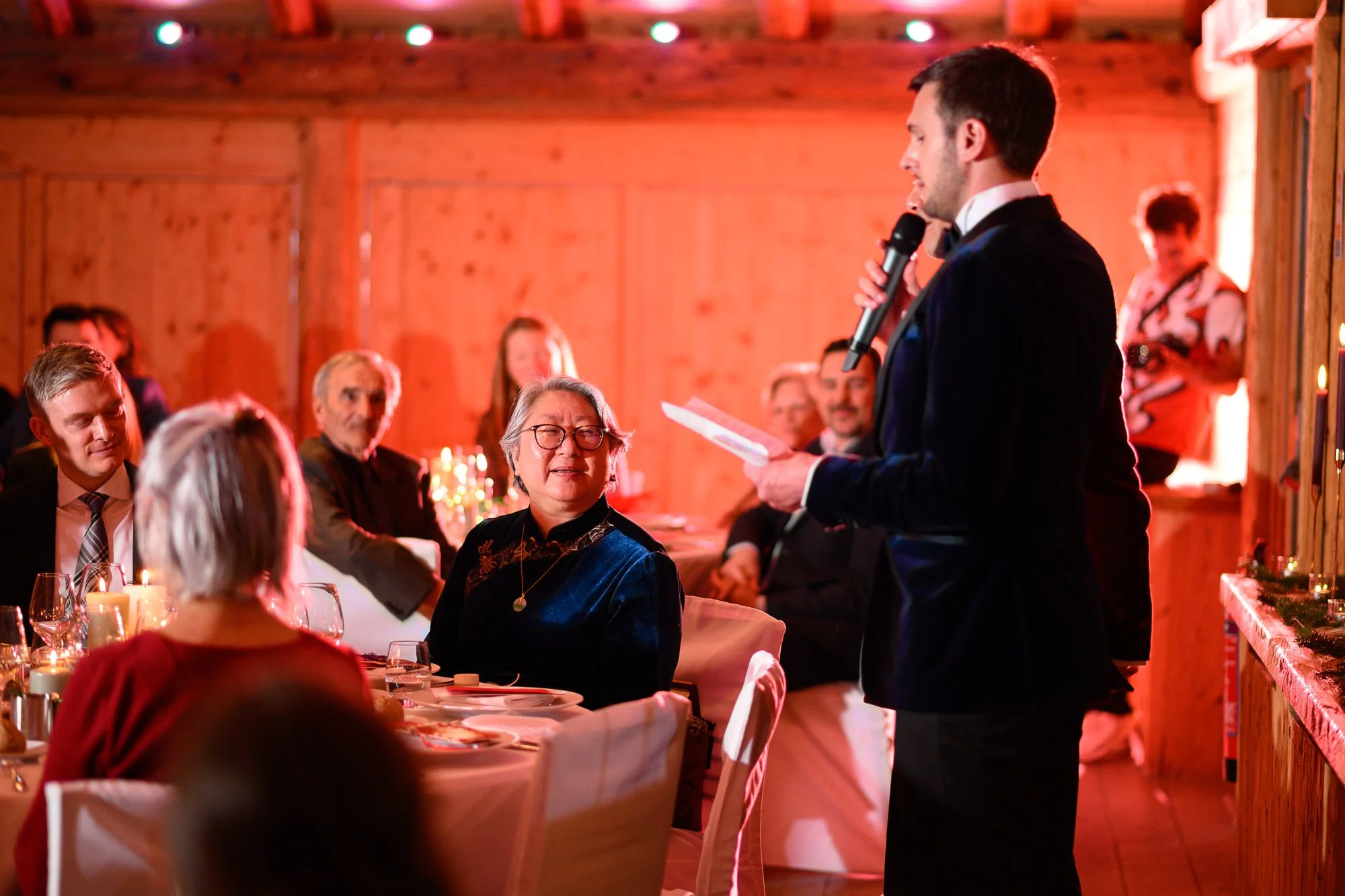 Un homme en costume noir parle dans un micro lors d'un événement dans une salle en bois, entouré de personnes assises à une table décorée, certaines souriantes et écoutant attentivement.
