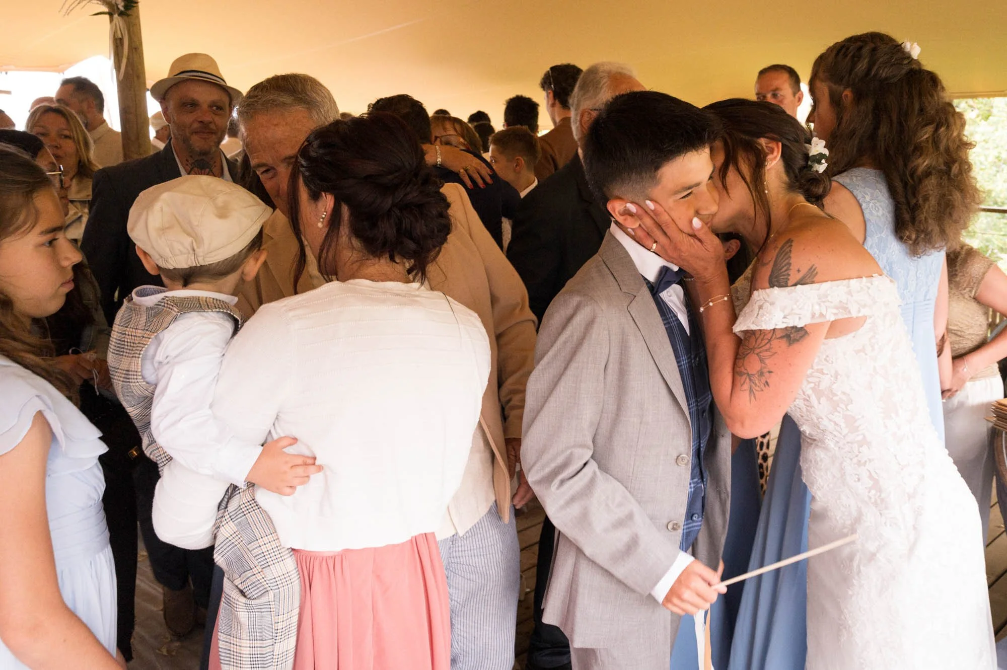 Une scène de mariage avec un groupe de personnes, dont un couple en train de s'embrasser, entouré de famille et amis dans une tente lors d'une célébration.