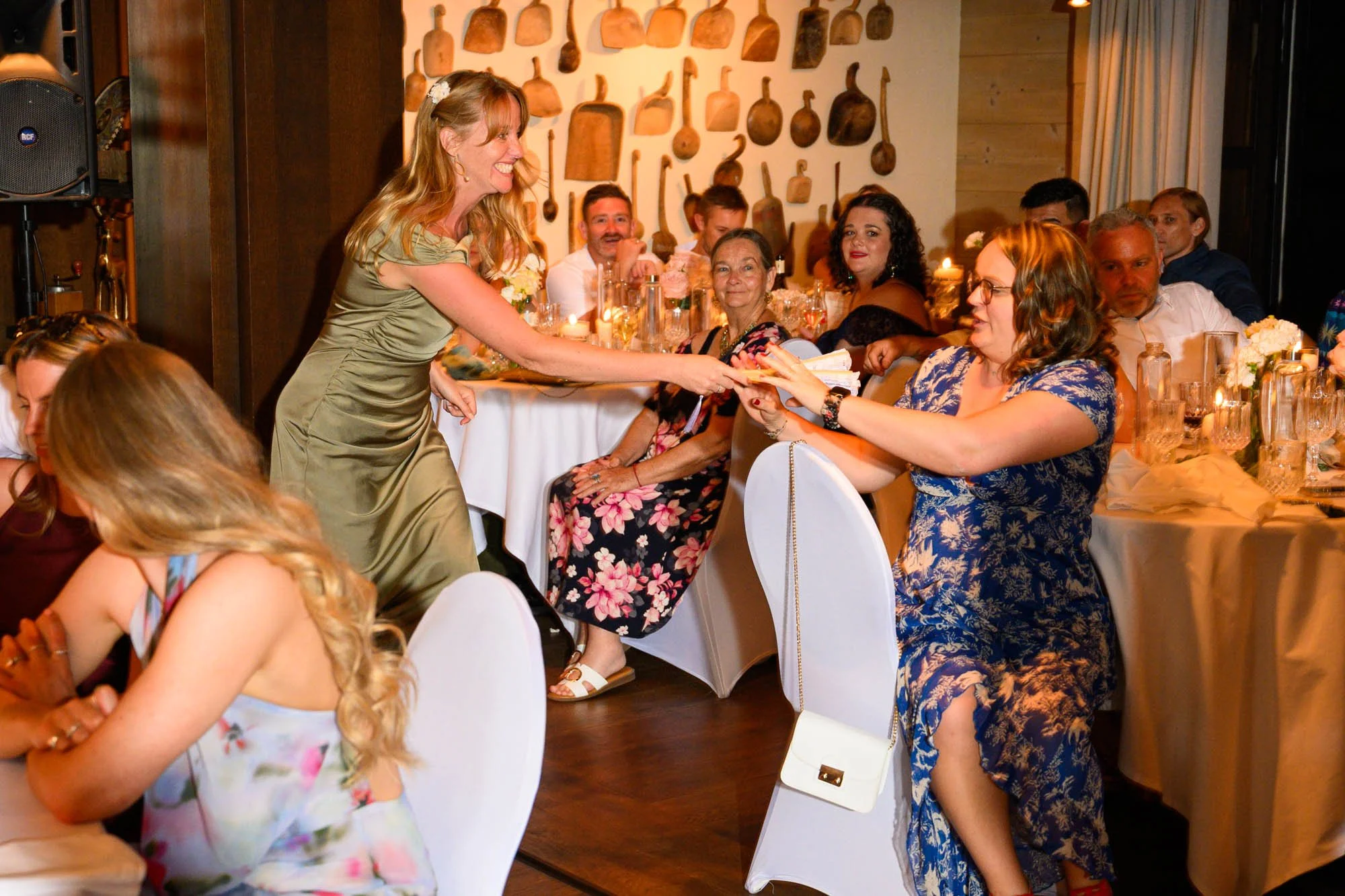Une femme en robe dorée échange un document avec une autre femme assise à une table lors d'une célébration, entourée de personnes souriantes dans un décor chaleureux avec des ustensiles en bois accrochés au mur.
