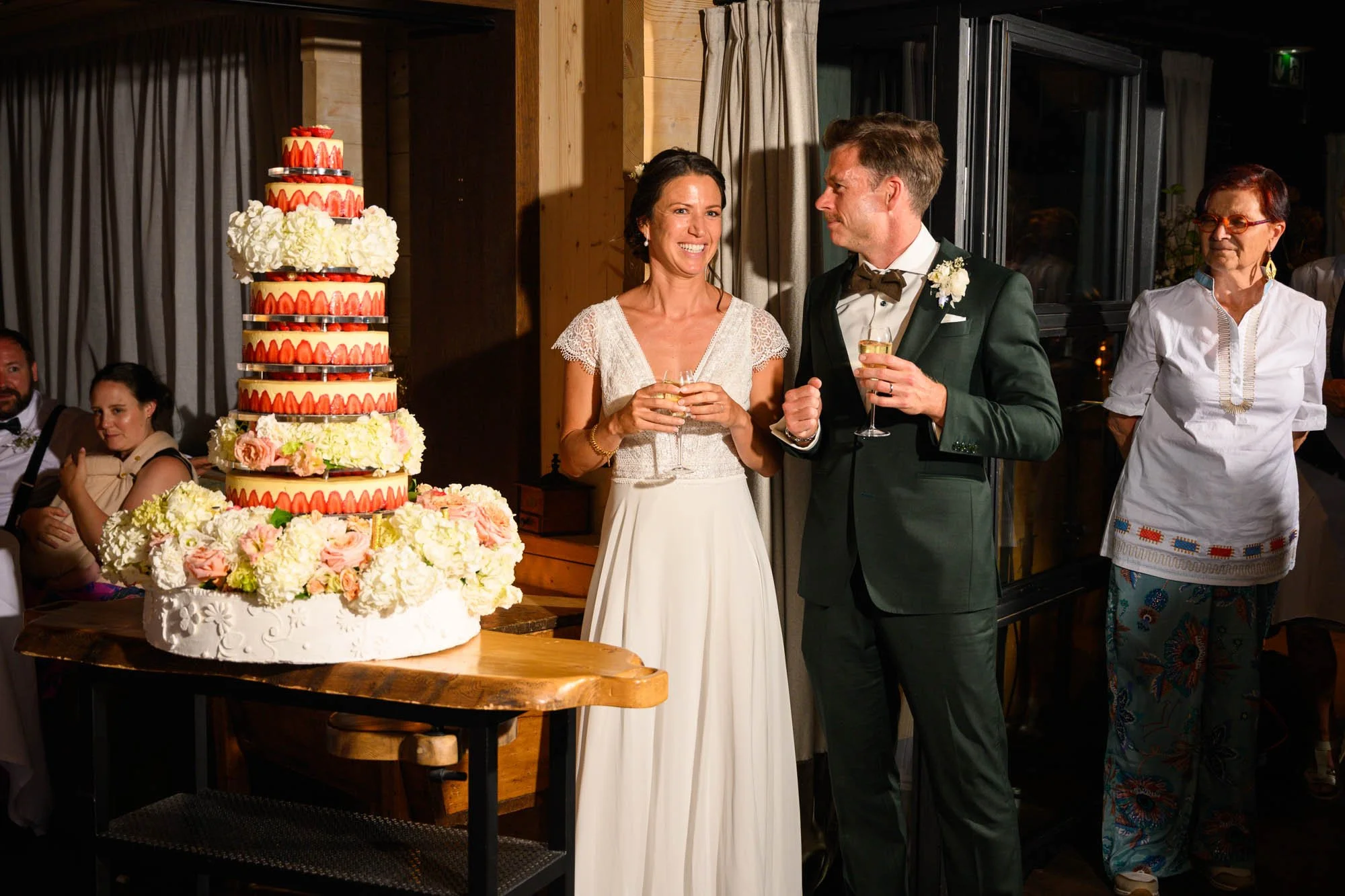 Un couple de mariés souriant lors de leur mariage, entourés de famille, avec un grand gâteau de mariage décoré de fleurs et de fraises à côté d'eux.