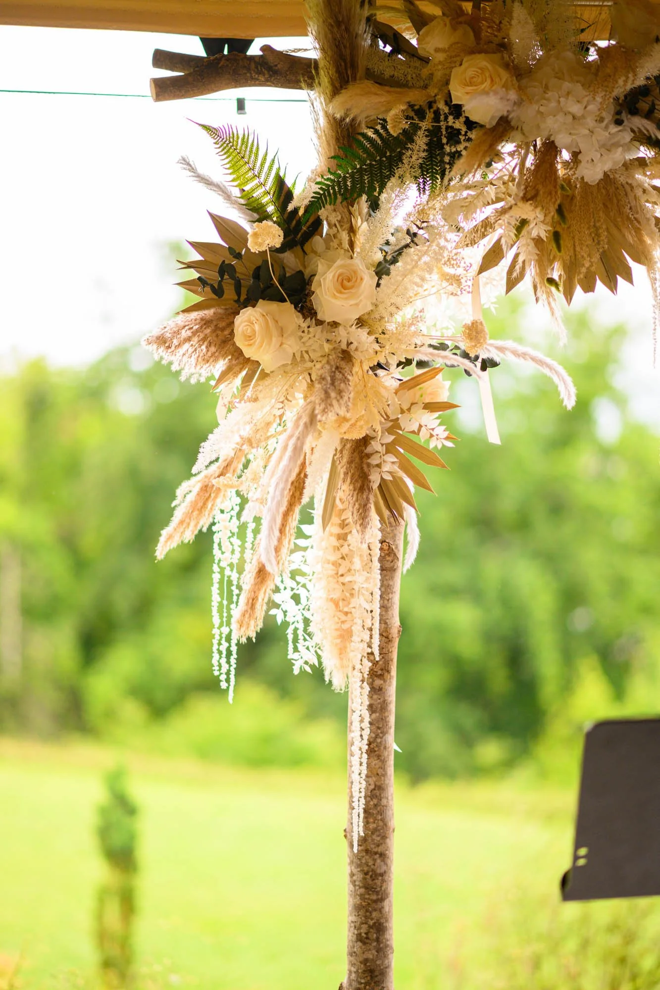 Une décoration florale suspendue composée de roses blanches, de fougères, de pampas blanches, et d'autres plantes décoratives, avec un fond naturel verdoyant.