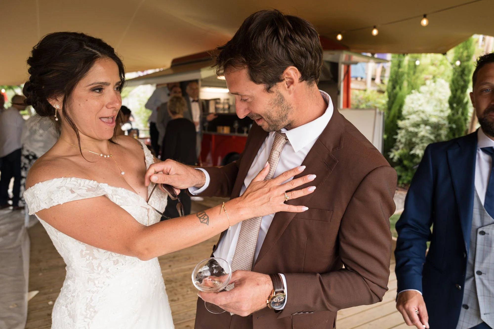 Un couple en vêtements de mariage en train de se faire une bague lors d'une cérémonie de mariage, en compagnie d'autres invités dans un lieu en plein air.