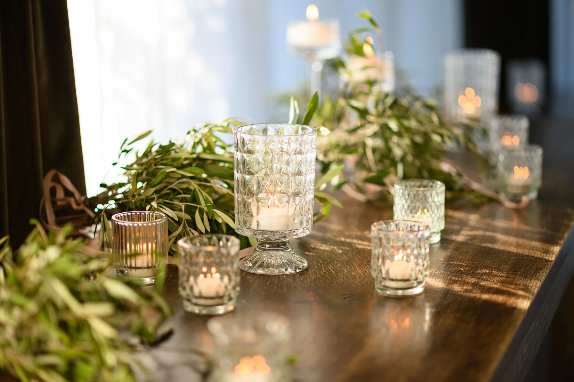 Bougies dans des verres en verre à motif posées sur une table en bois, avec des feuillages verts en décoration.
