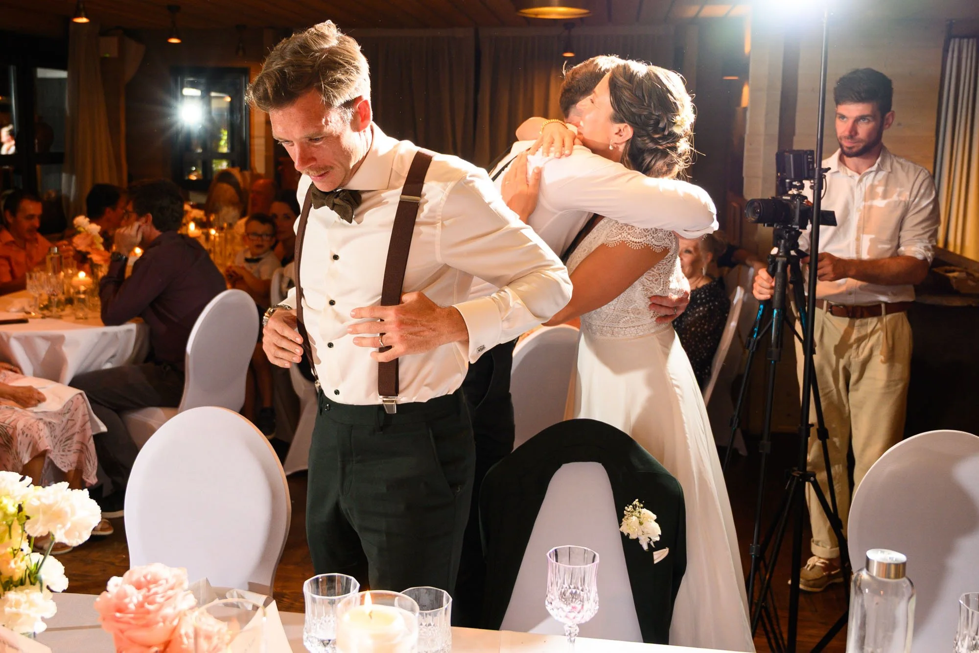 Un homme en smoking blanc avec improbateur et une femme en robe blanche de mariage en train de s'embrasser lors d'une réception. Un autre homme avec une caméra observe la scène dans une pièce chaleureuse.