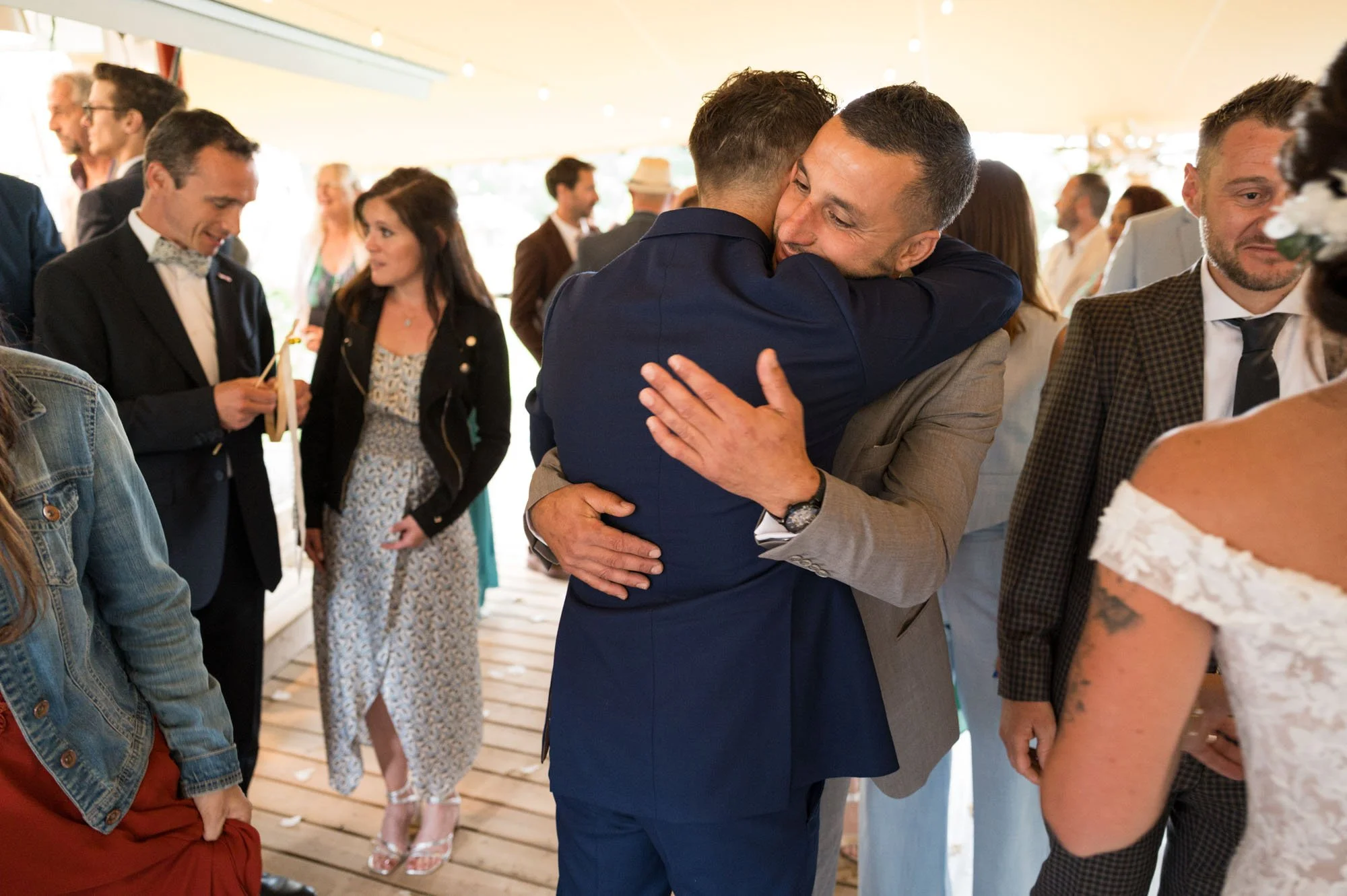 Deux hommes s'embrassent lors d'une célébration, entourés d'autres personnes en tenues habillées.