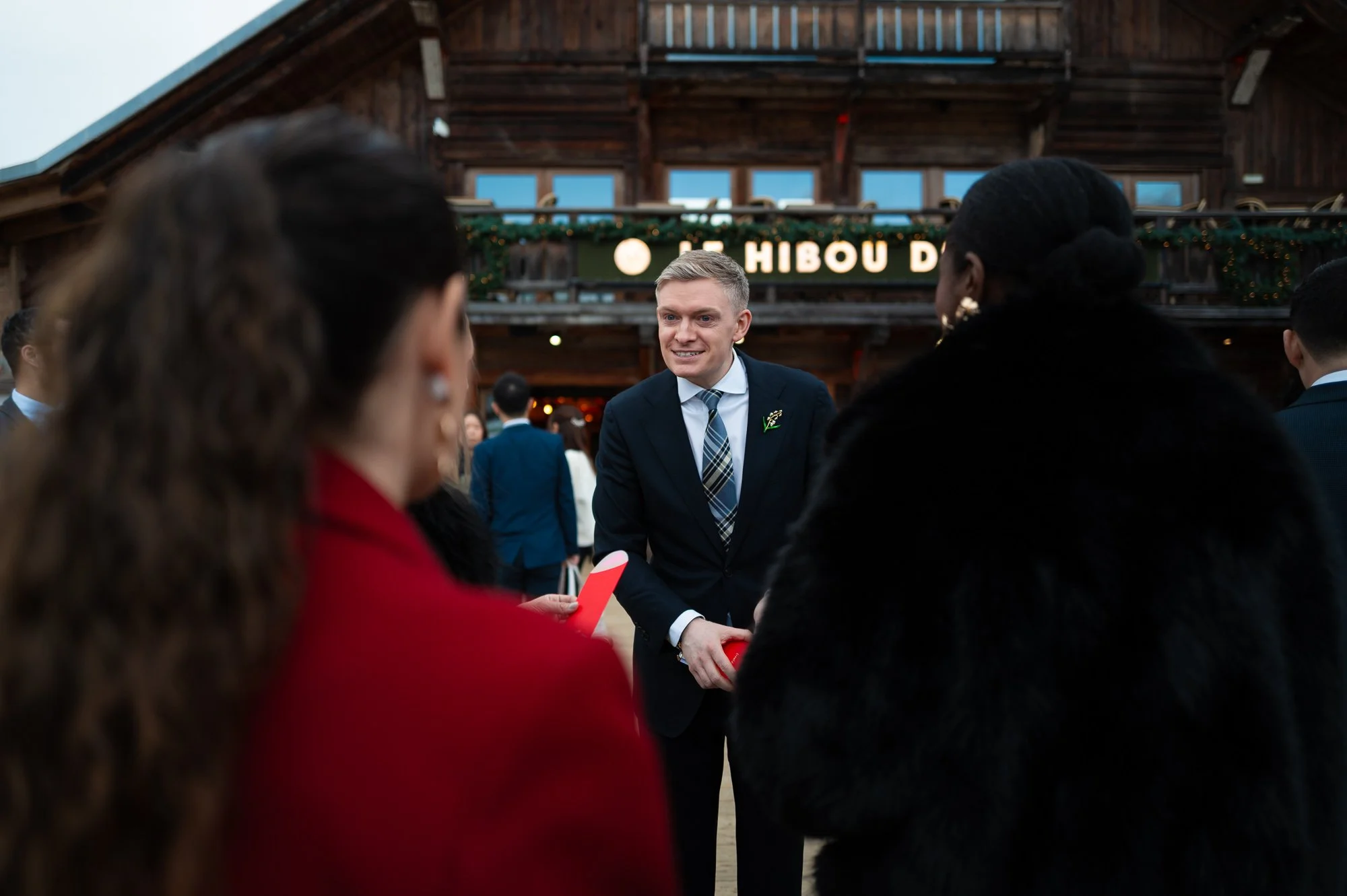 Un homme en costume parle avec plusieurs femmes lors d'un événement en intérieur, avec un fond en bois et un panneau lumineux avec le texte 'Hibou D' dans une ambiance festive.