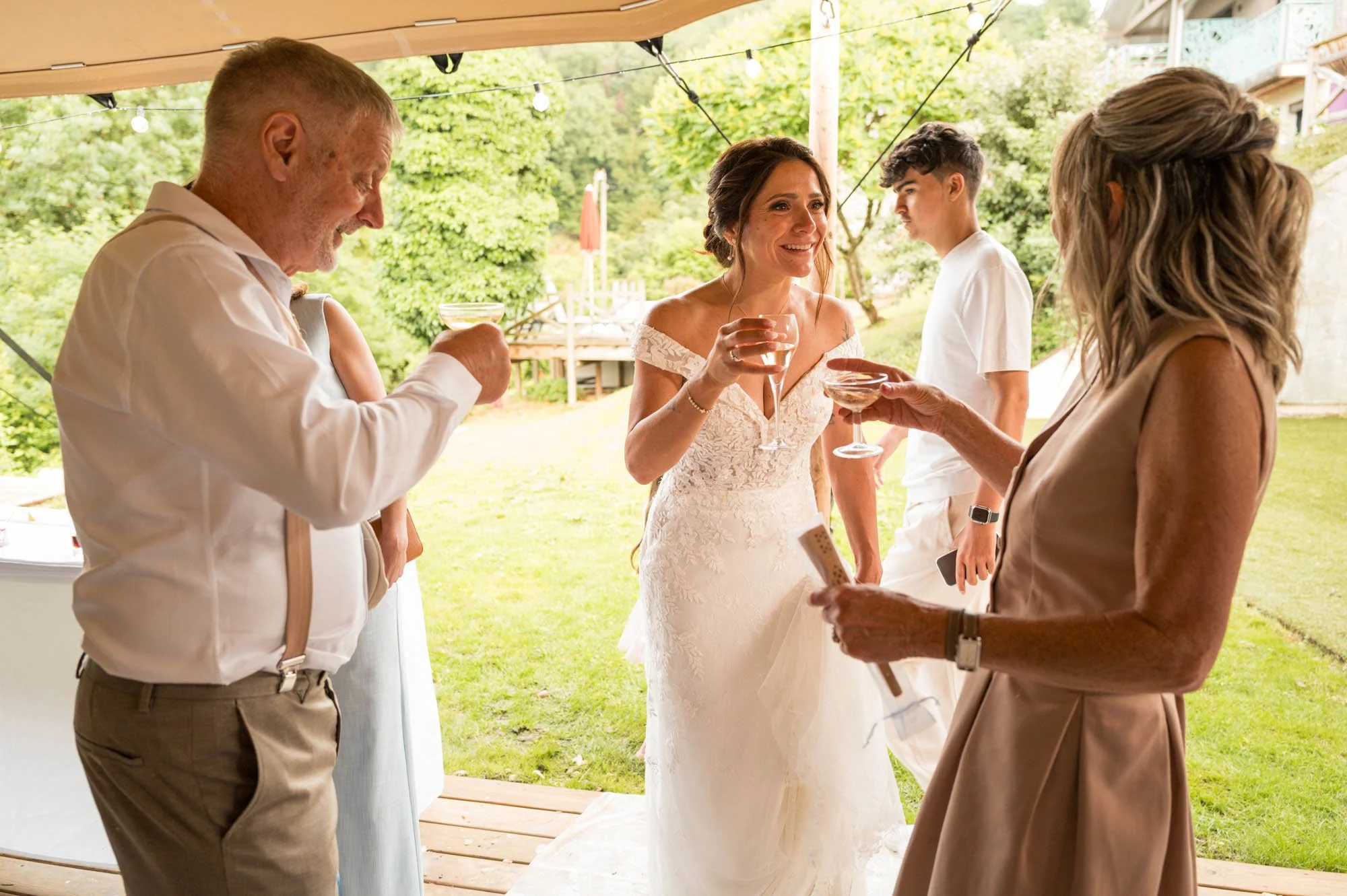 Groupe de personnes lors d'une fête en extérieur, avec une femme en robe de mariée au centre, tenant un verre, entourée d'autres adultes qui se tiennent et parlent dans une ambiance chaleureuse avec des lumières suspendues.