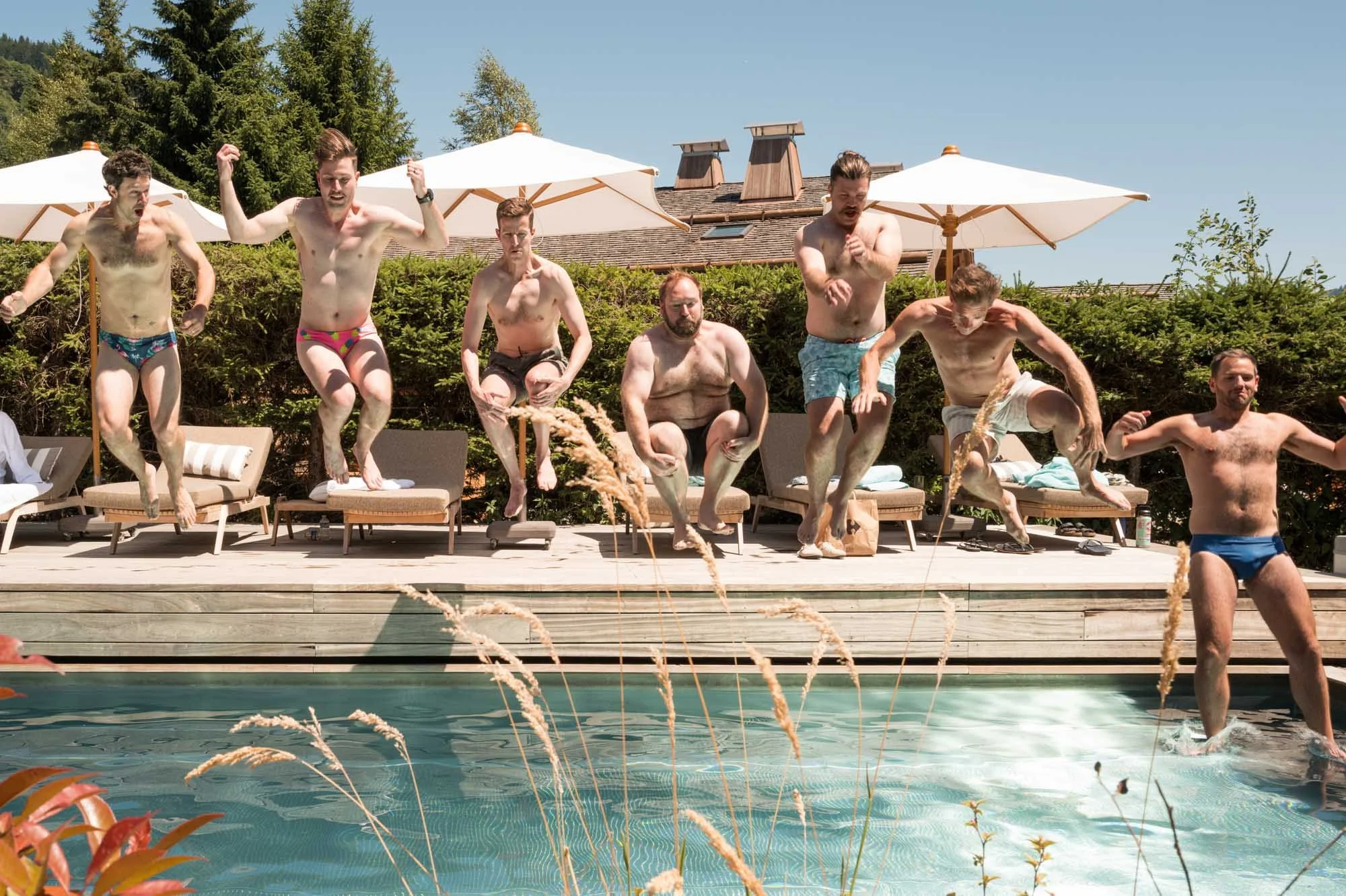 Groupe d'hommes en maillots de bain sautant dans une piscine lors d'une activité aquatique en plein air, avec parasols et végétation en arrière-plan.
