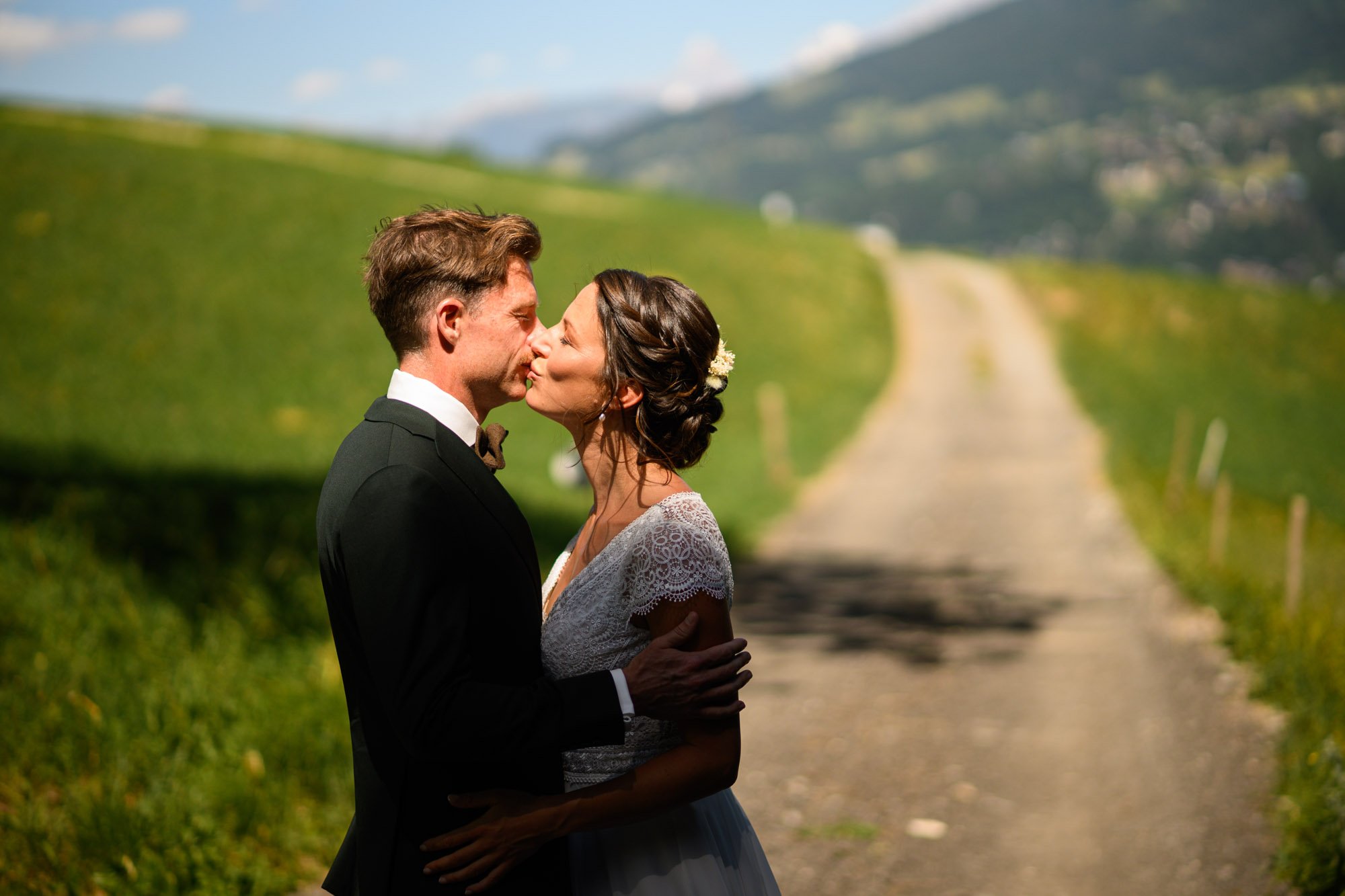 Un couple en robe de mariage s'embrasse sur un chemin de campagne vert, avec des montagnes en fond.