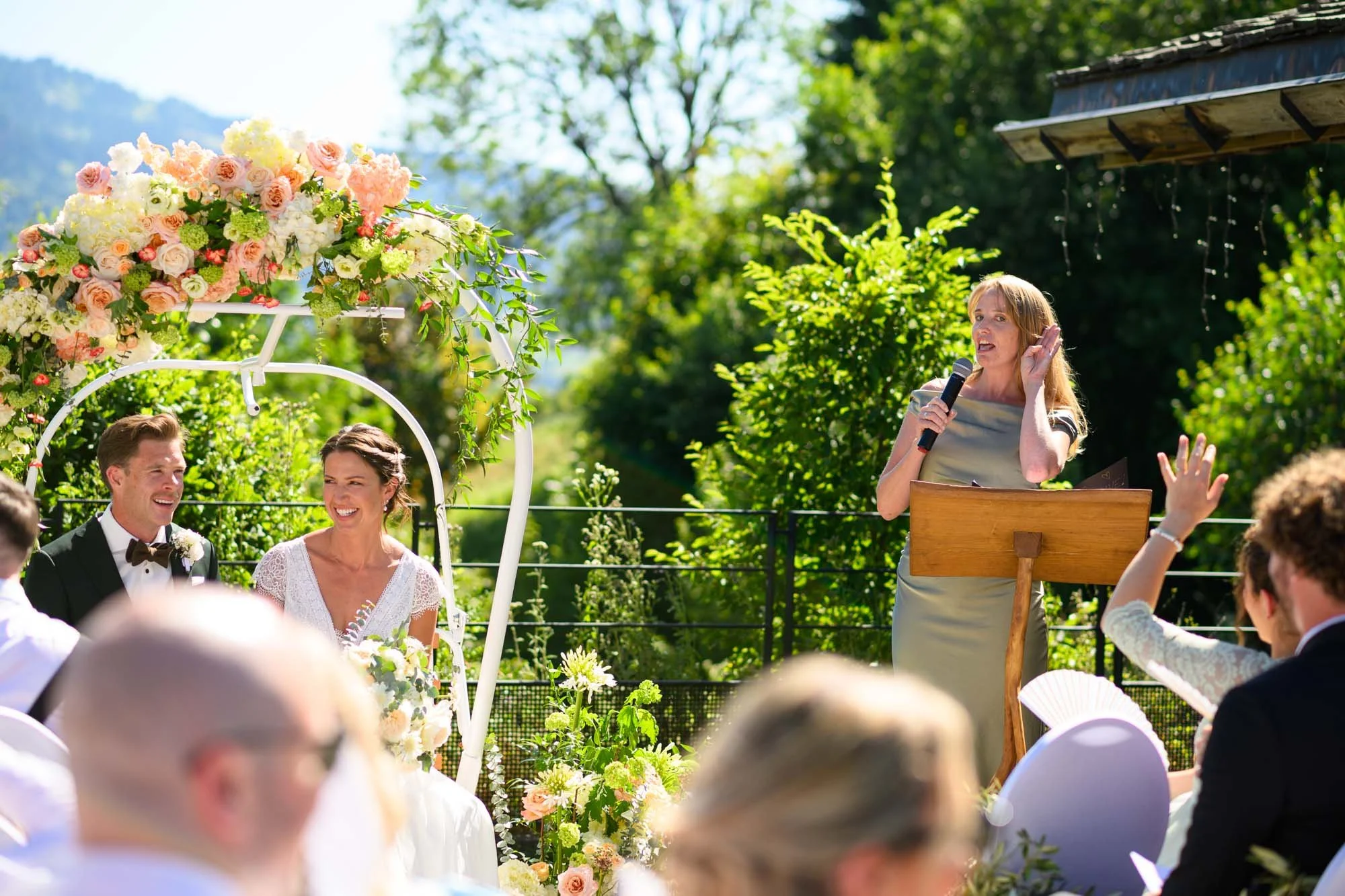 Cérémonie de mariage en plein air avec un couple assis sous une arche décorée de fleurs, une officiant parlant dans un micro, entouré d'invités. Nature et ciel en arrière-plan.